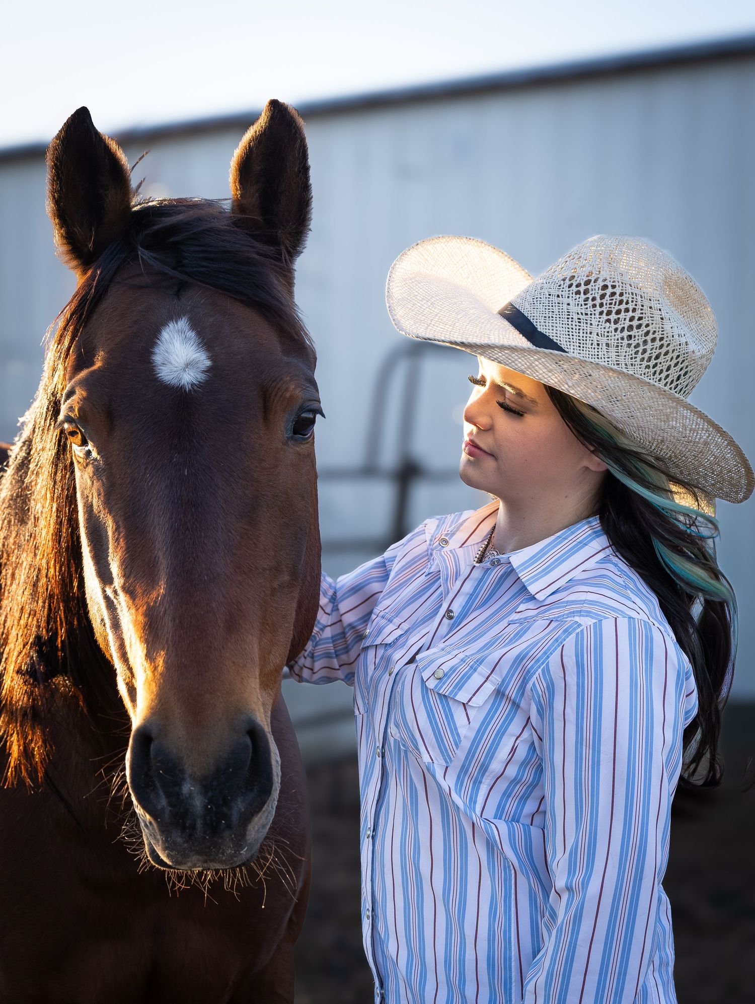 Young woman in cowboy hat and striped western shirt gently touches a brown horse near a barn.