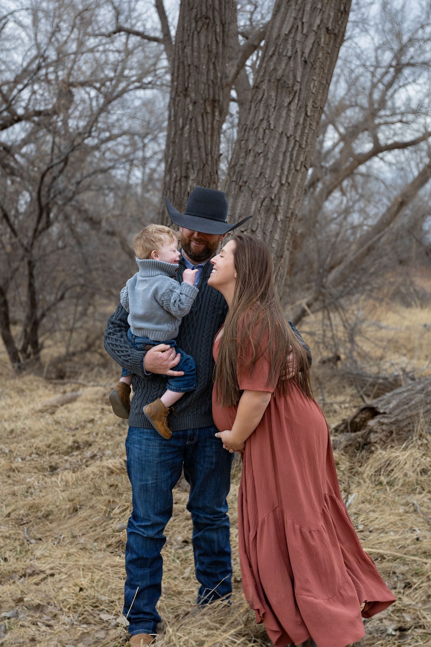 Man in cowboy hat holds toddler while smiling at pregnant woman in rust dress near large tree.
