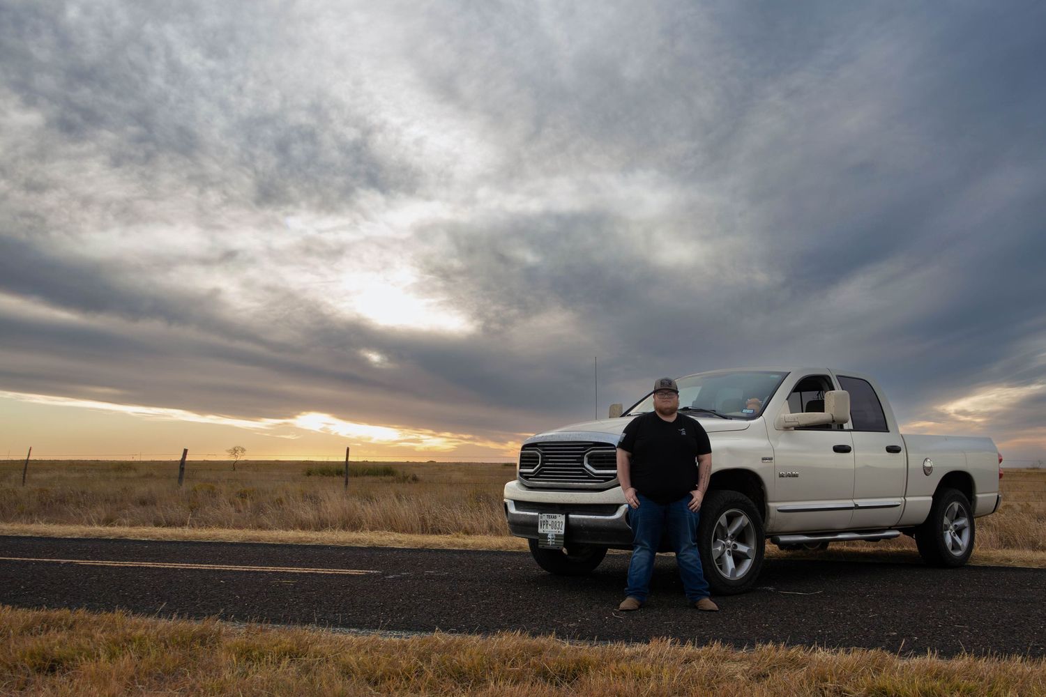 Man standing beside white Dodge Ram pickup truck on rural road under dramatic cloudy sunset sky.