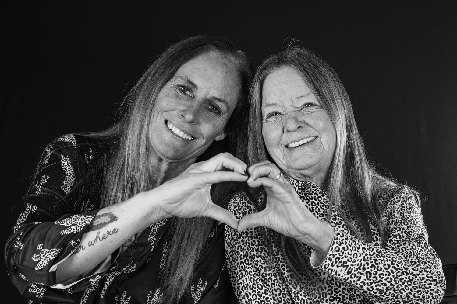 Two smiling women making heart shape with hands together in black and white portrait photo.