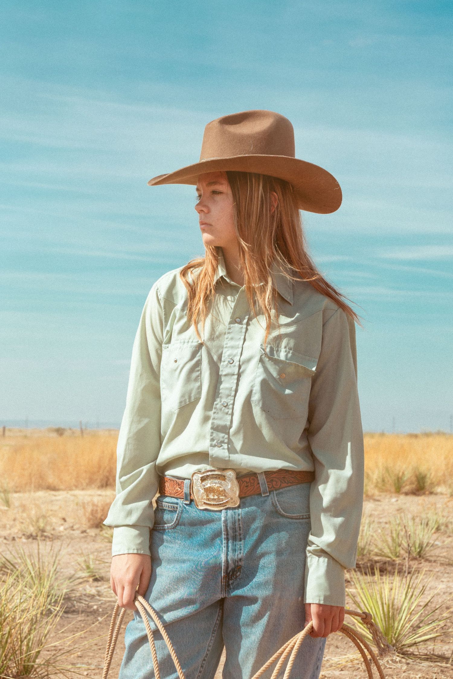 Young cowgirl in tan hat and light shirt holding lasso in dry desert grassland under blue sky.