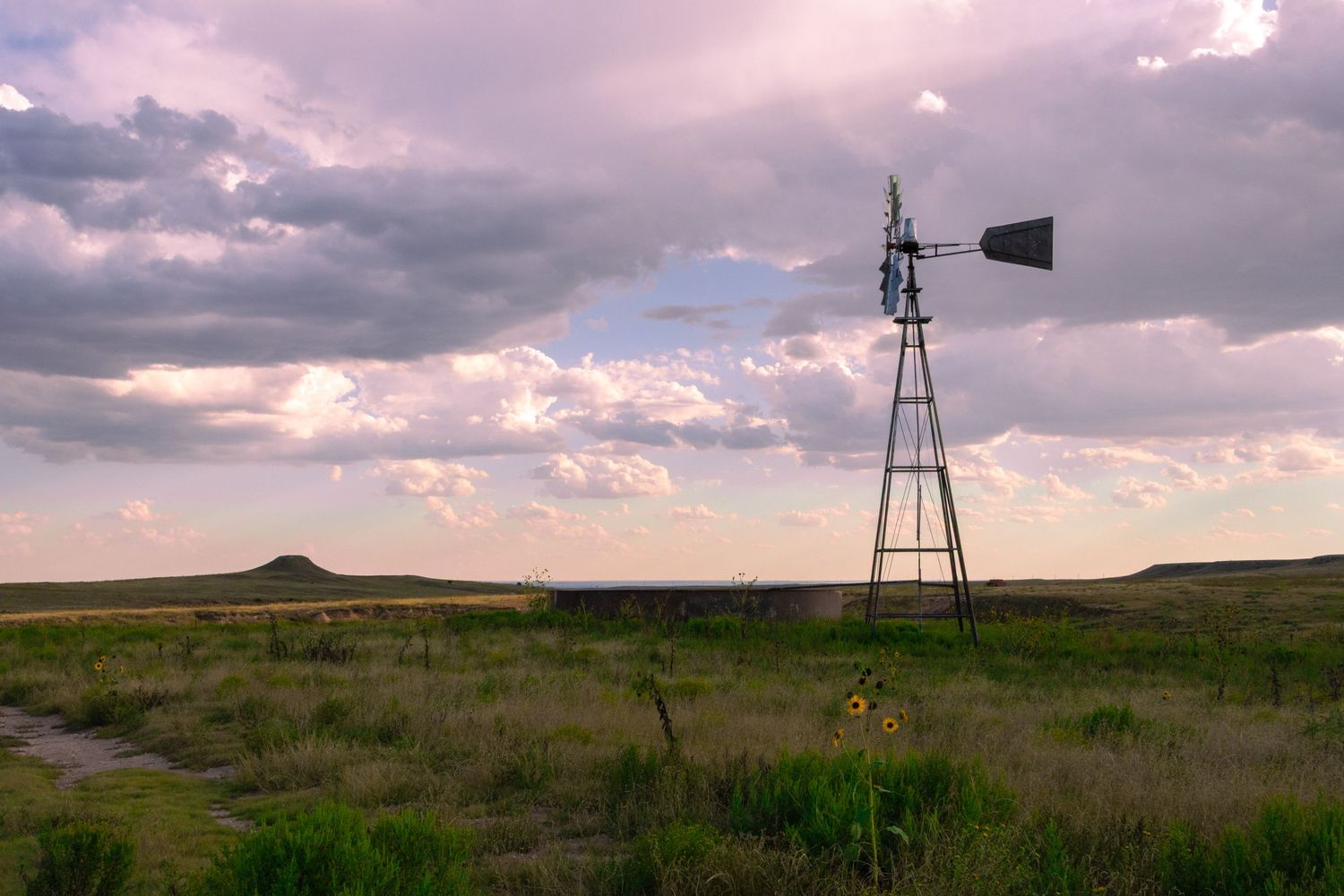 Vintage windmill standing in green prairie grassland under dramatic cloudy sky at dusk.
