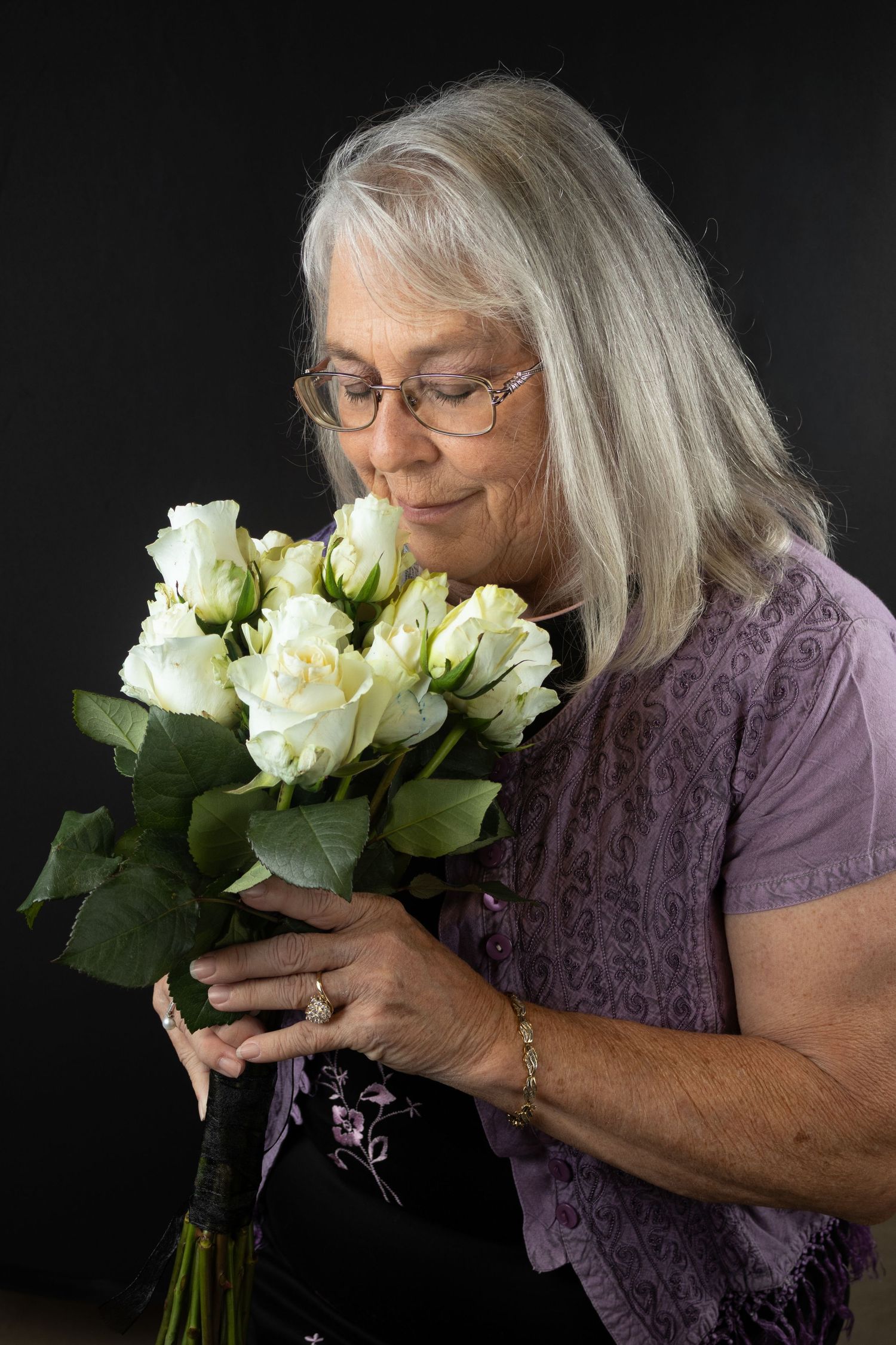 Older woman with gray hair and glasses smelling bouquet of white roses on dark background.