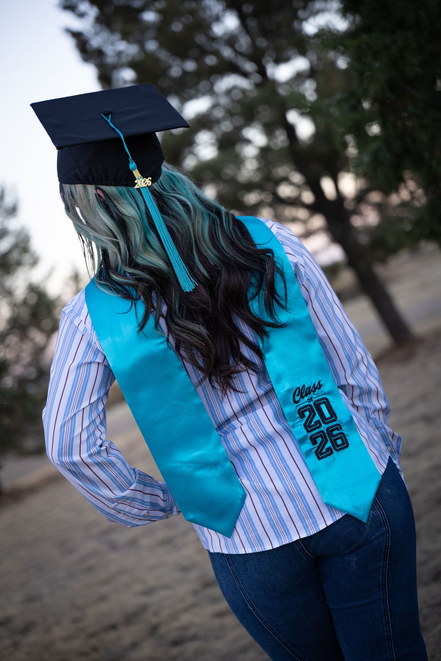 Back view of graduate with teal-highlighted hair wearing Class of 2025 turquoise stole and black cap outdoors.