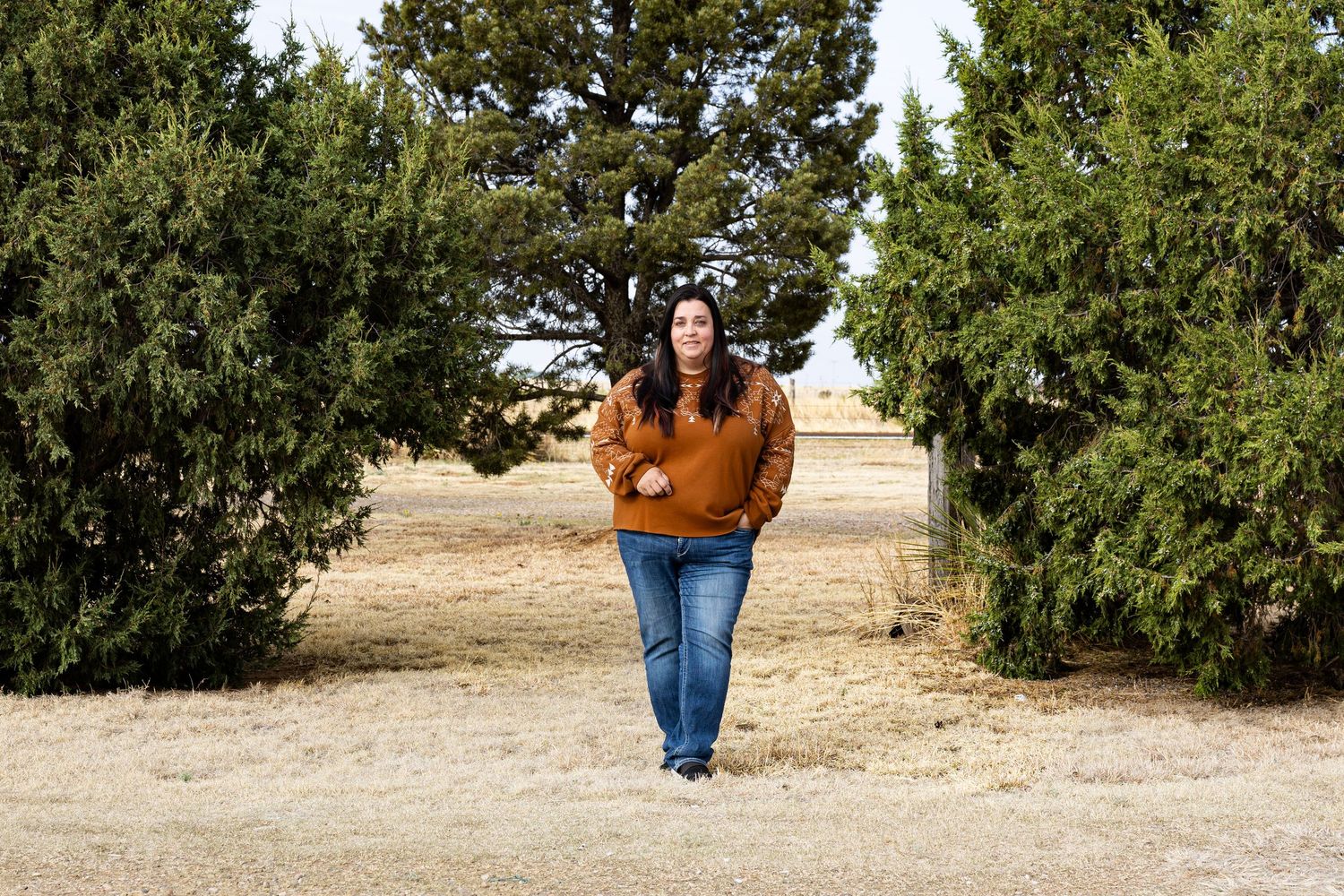 Woman in orange sweater walking between cedar trees on dry grassland.