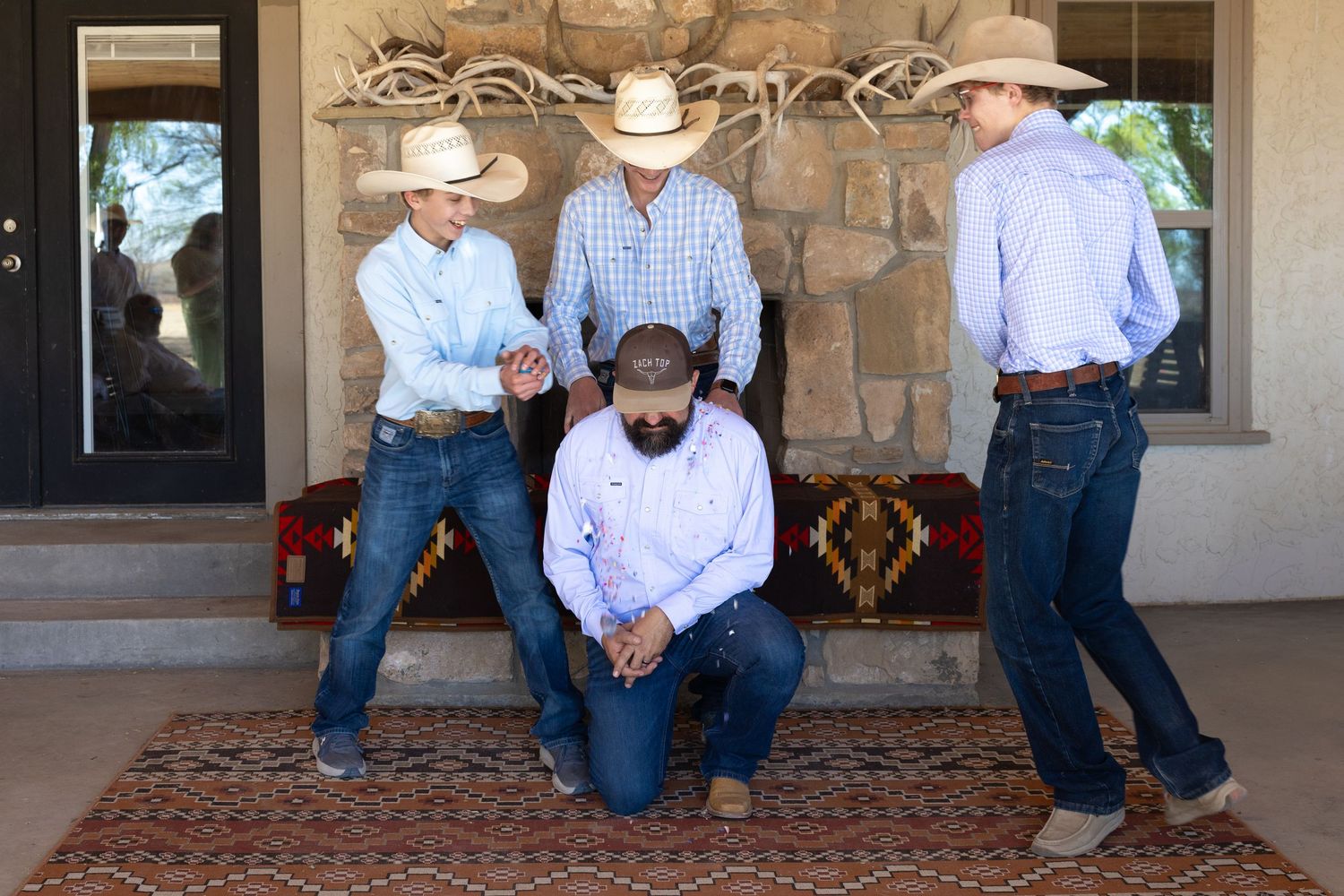 Four men in western wear and cowboy hats gather on a ranch porch with a colorful southwestern rug.
