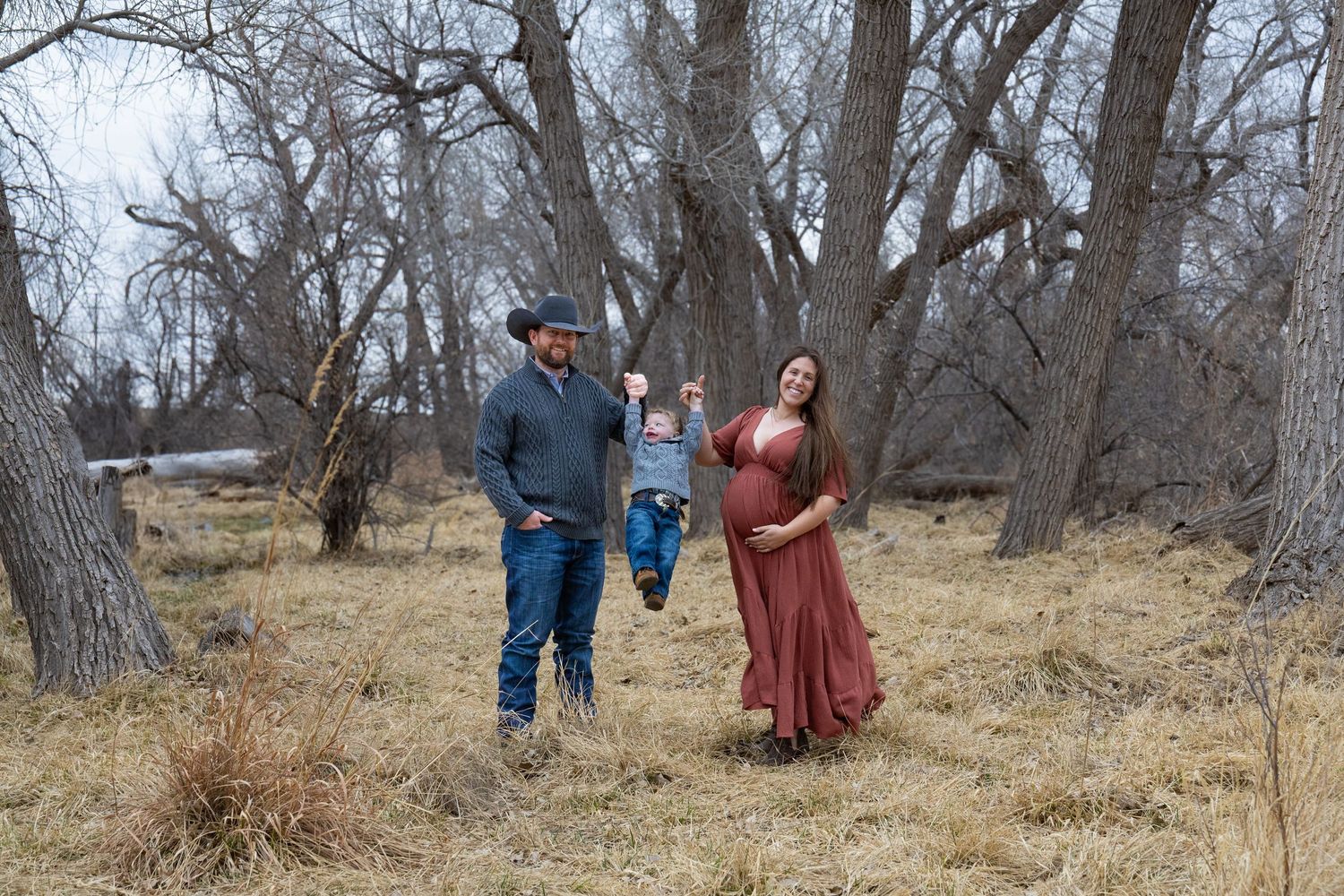 Family portrait with father lifting toddler beside pregnant mother in rust dress among bare trees.