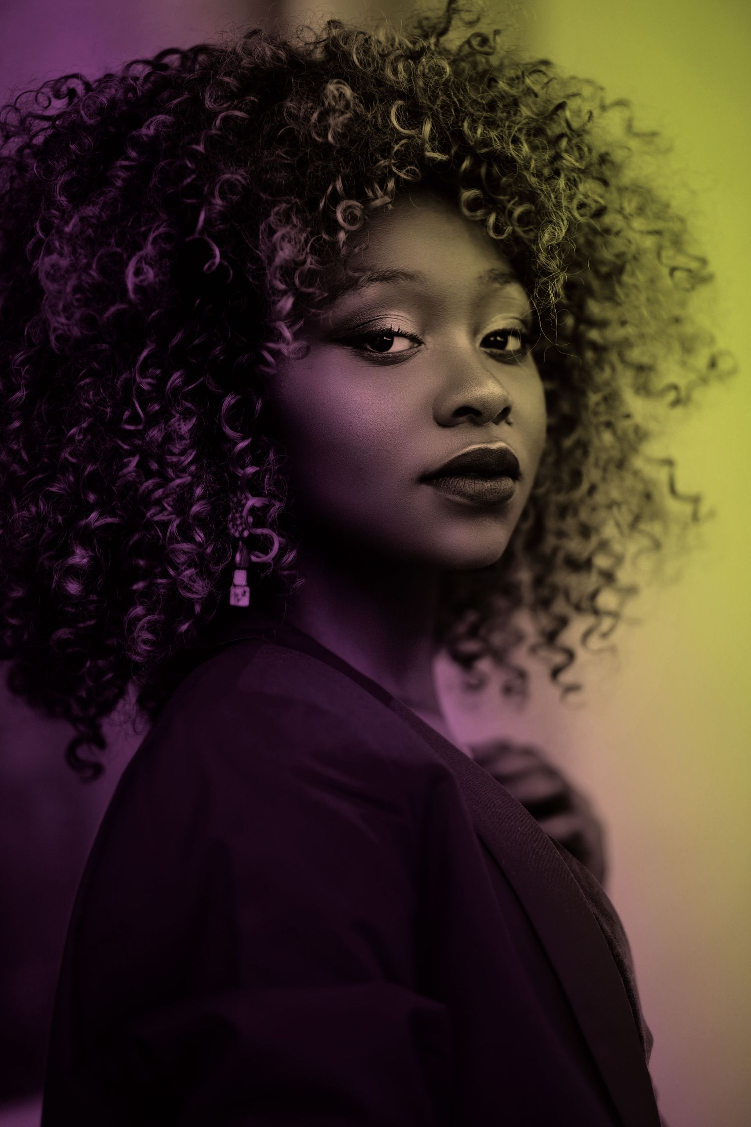 Portrait of a young Black woman with curly hair under striking purple and yellow neon lighting.