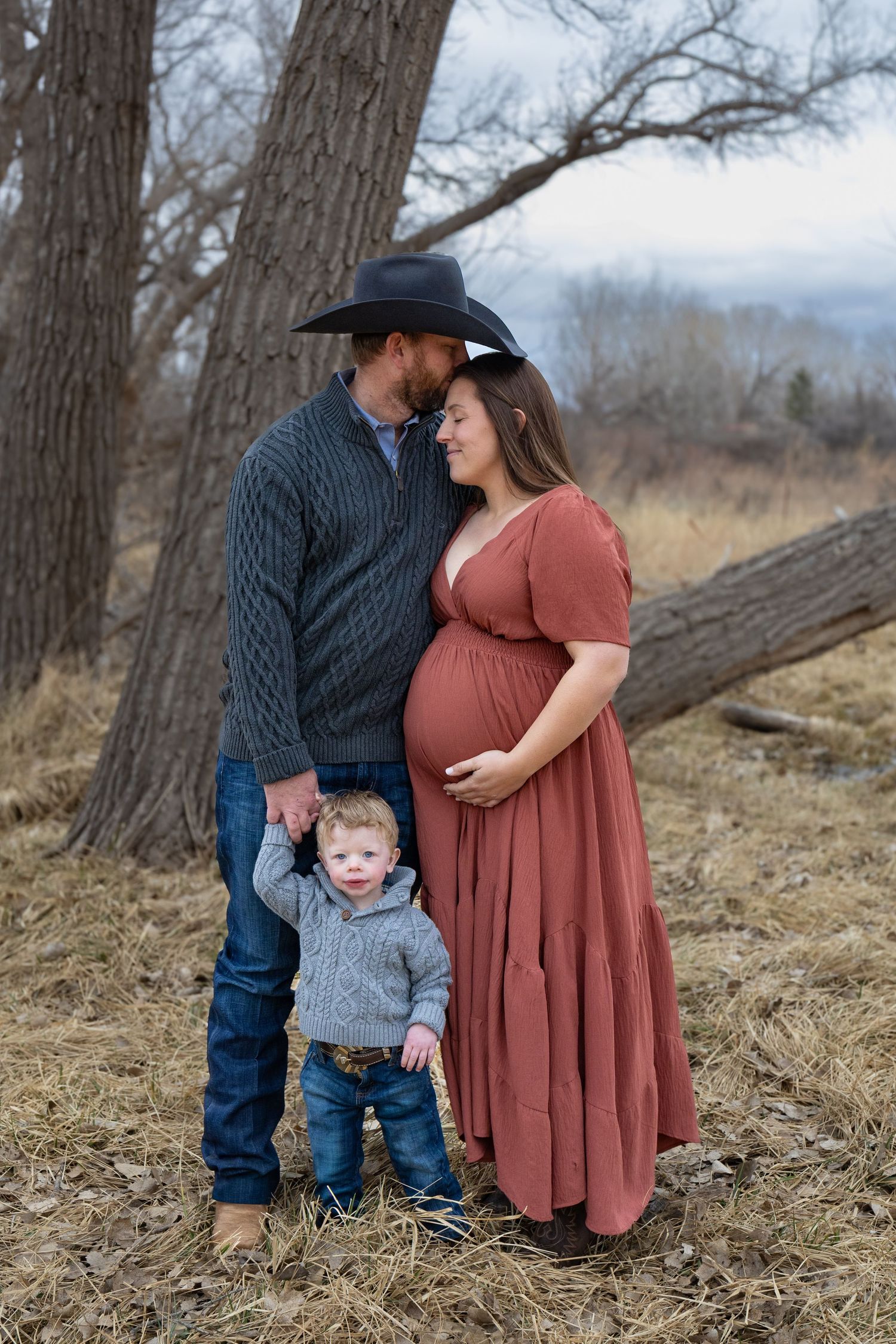 Couple with cowboy hat dad and pregnant mom in rust dress share tender moment with toddler.
