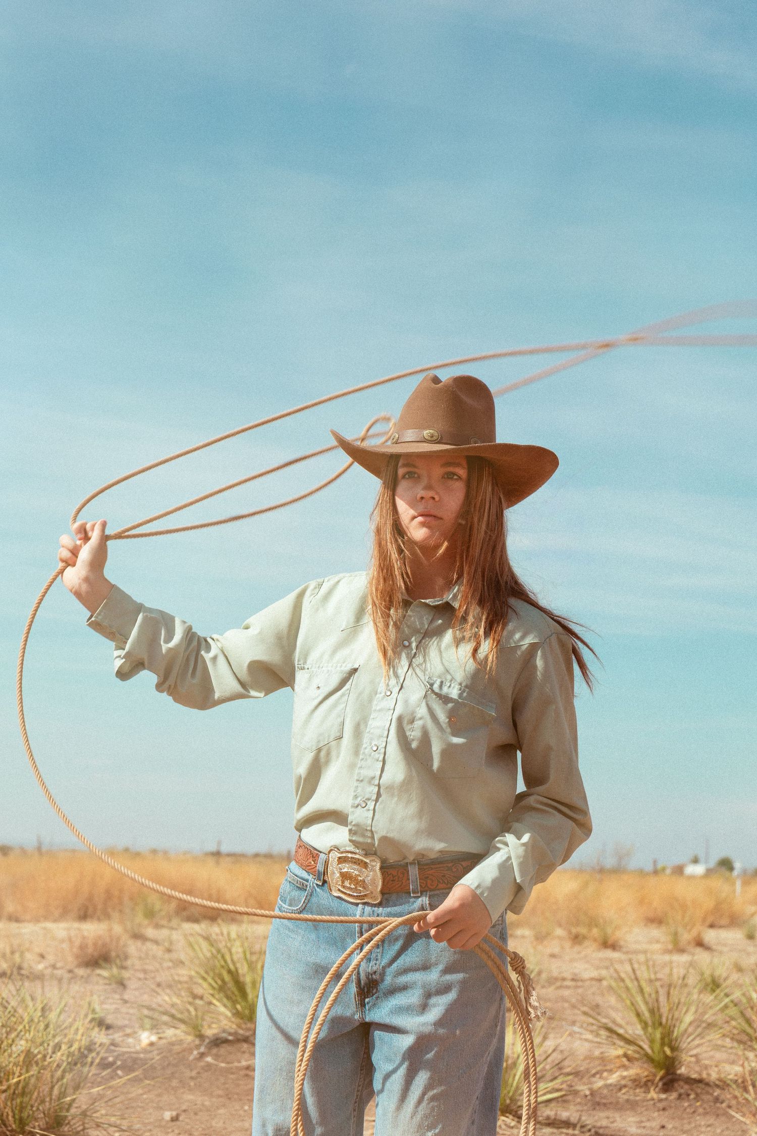 Young cowgirl in wide-brim hat swinging lasso rope in arid desert landscape under partly cloudy sky.