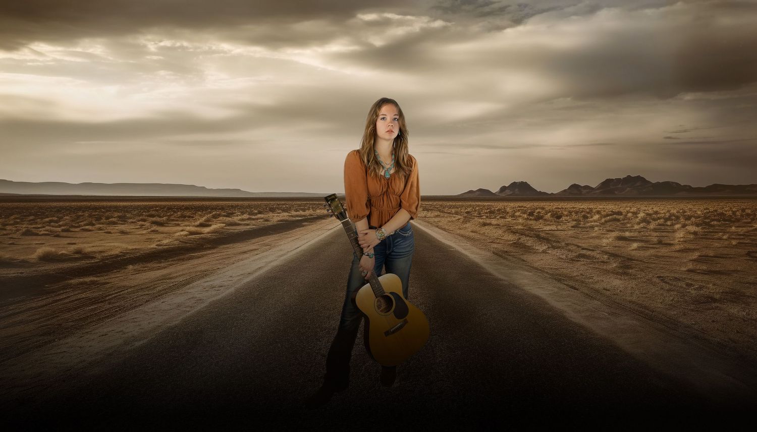Woman with acoustic guitar standing on empty desert highway under dramatic sky.