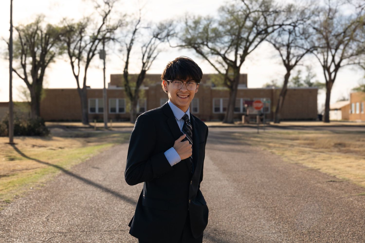 Smiling young man in a dark suit carrying a bag, standing on a tree-lined school pathway outdoors.