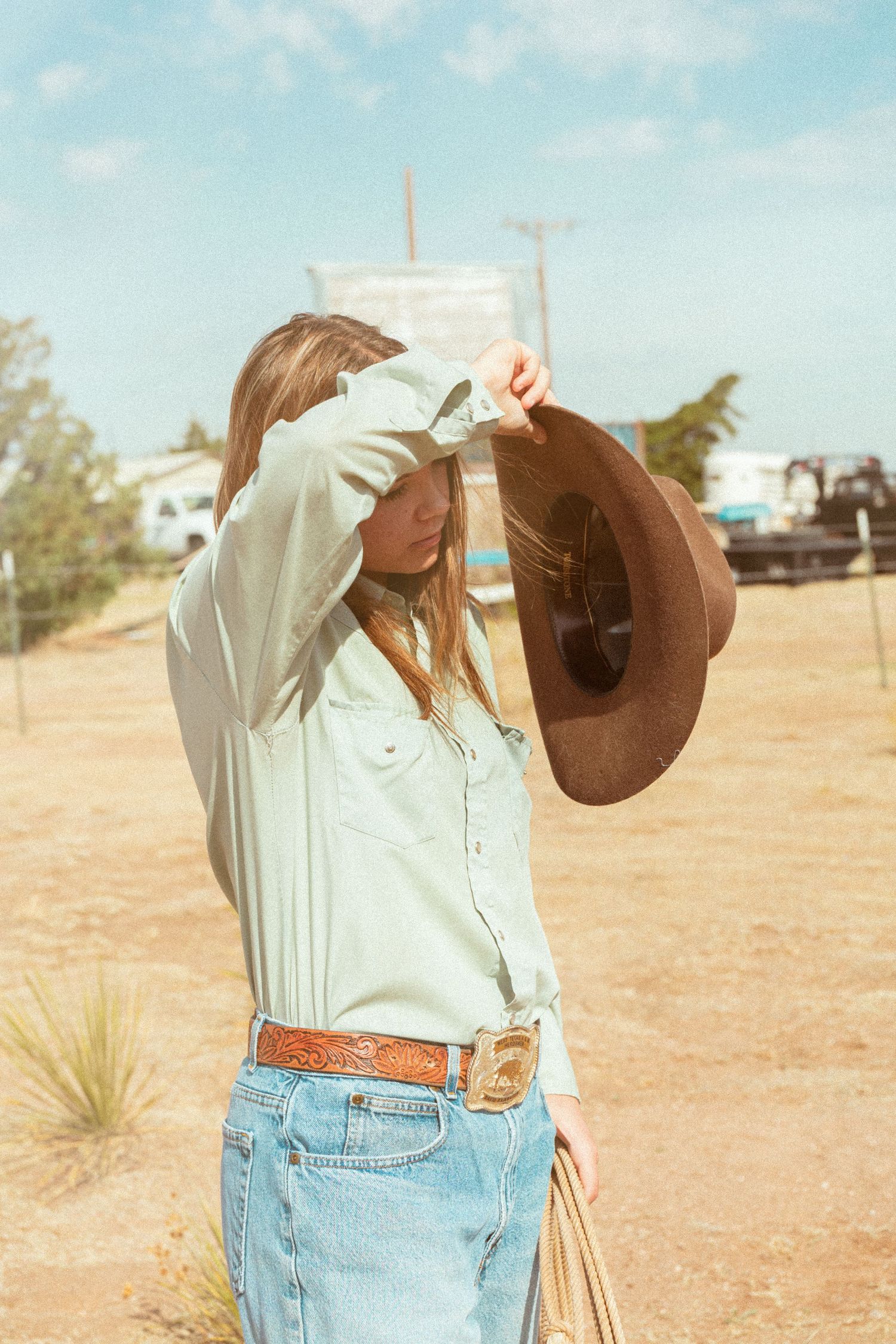 Young woman lifting brown cowboy hat above her head in sunny desert ranch setting with trailers.