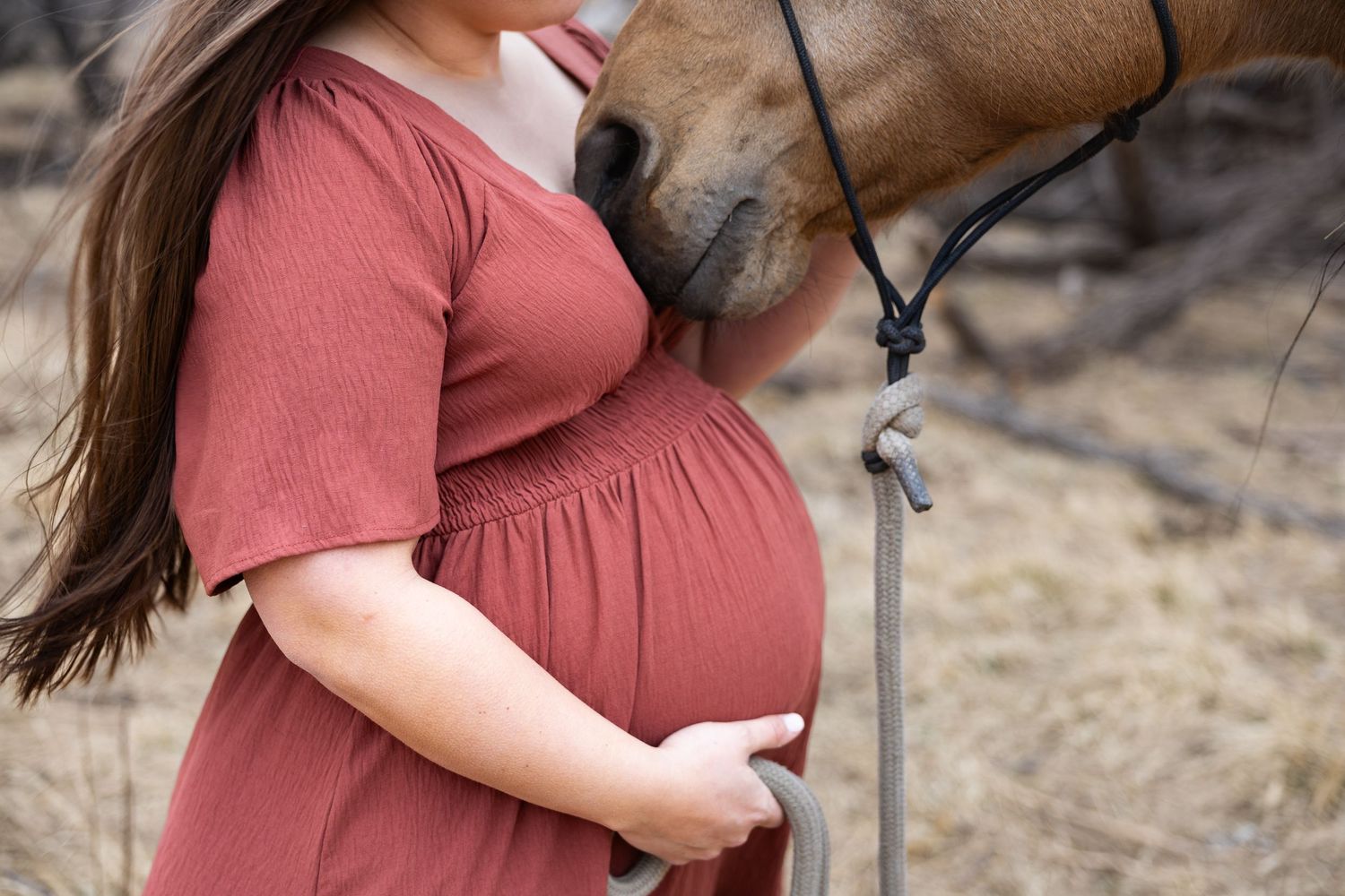 Pregnant woman in rust dress cradling baby bump while horse nuzzles her from behind.