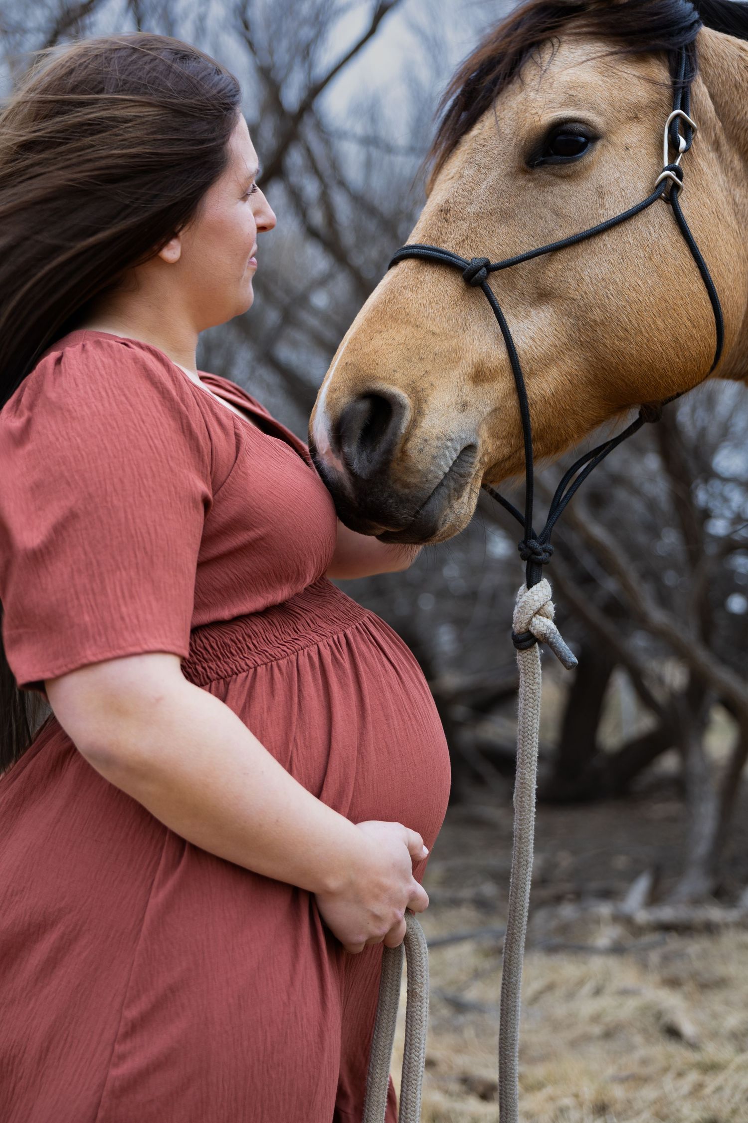 Pregnant woman in rust dress stands beside a tan horse with black bridle in an outdoor setting.