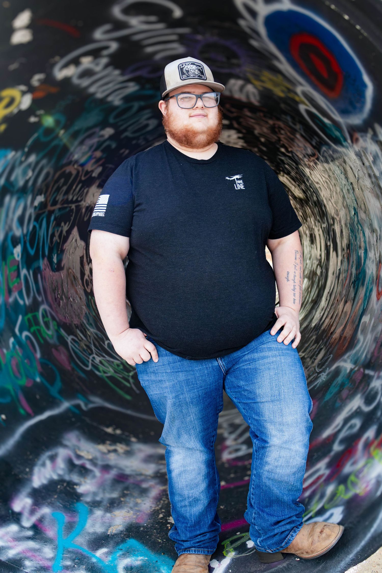 Young man in black t-shirt and cap posing inside graffiti-covered tunnel with fisheye perspective effect.