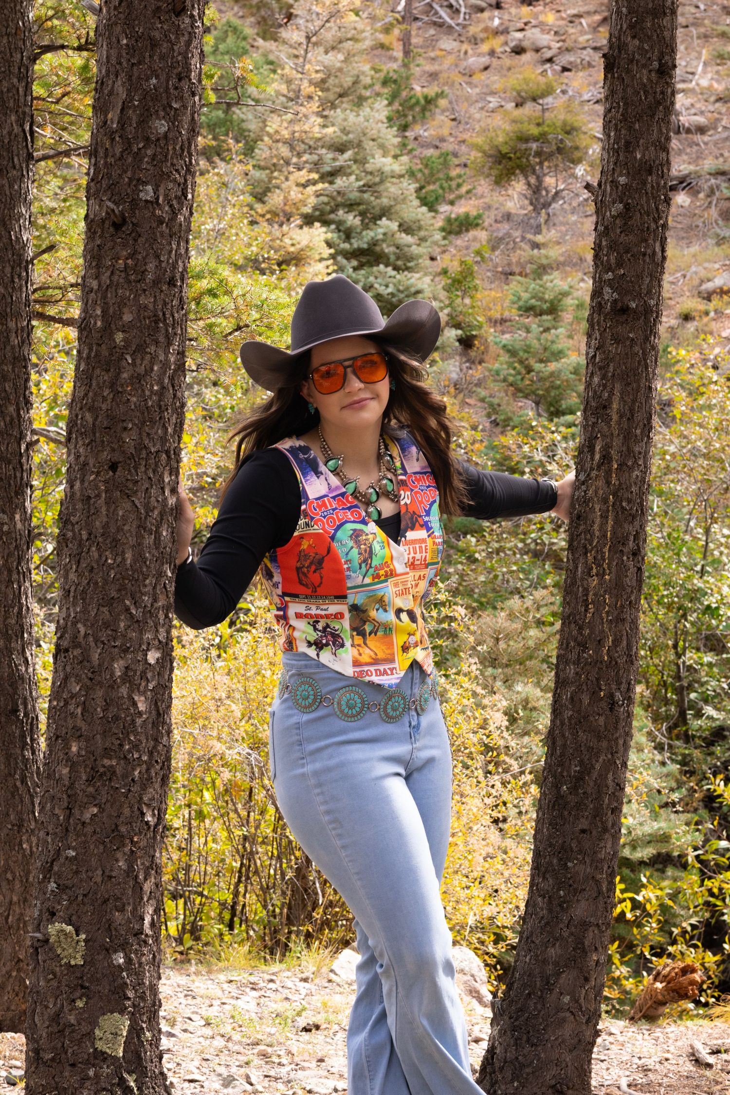 Young woman in cowboy hat, orange sunglasses, colorful vest, and flare jeans poses between trees outdoors.