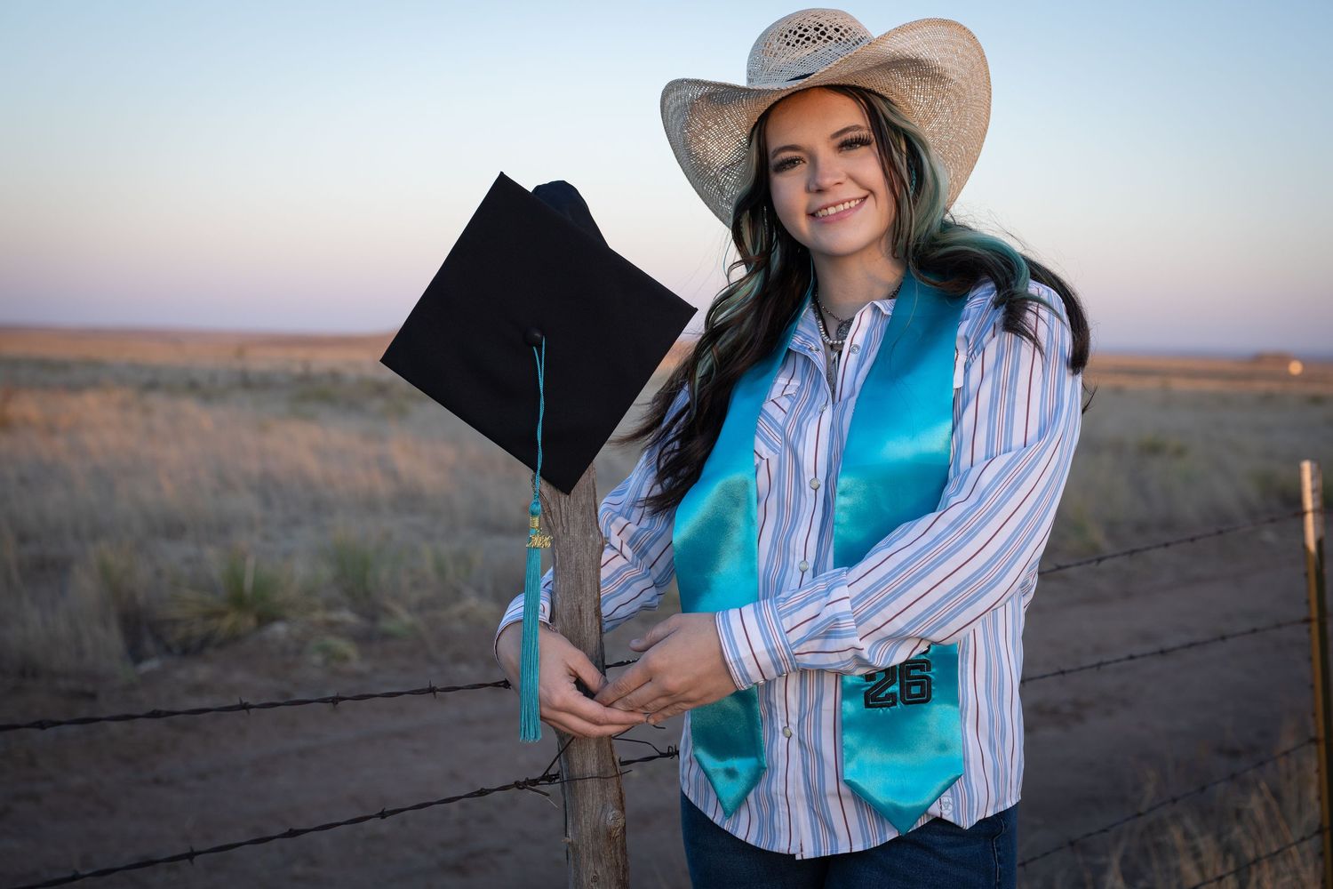 Smiling graduate in cowboy hat and turquoise sash holds black cap beside wooden fence post in open field.