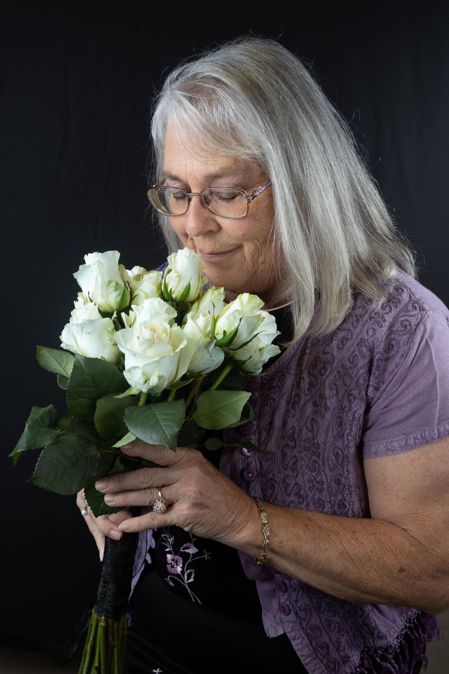 Elderly woman with gray hair and glasses smelling bouquet of white roses against dark background.