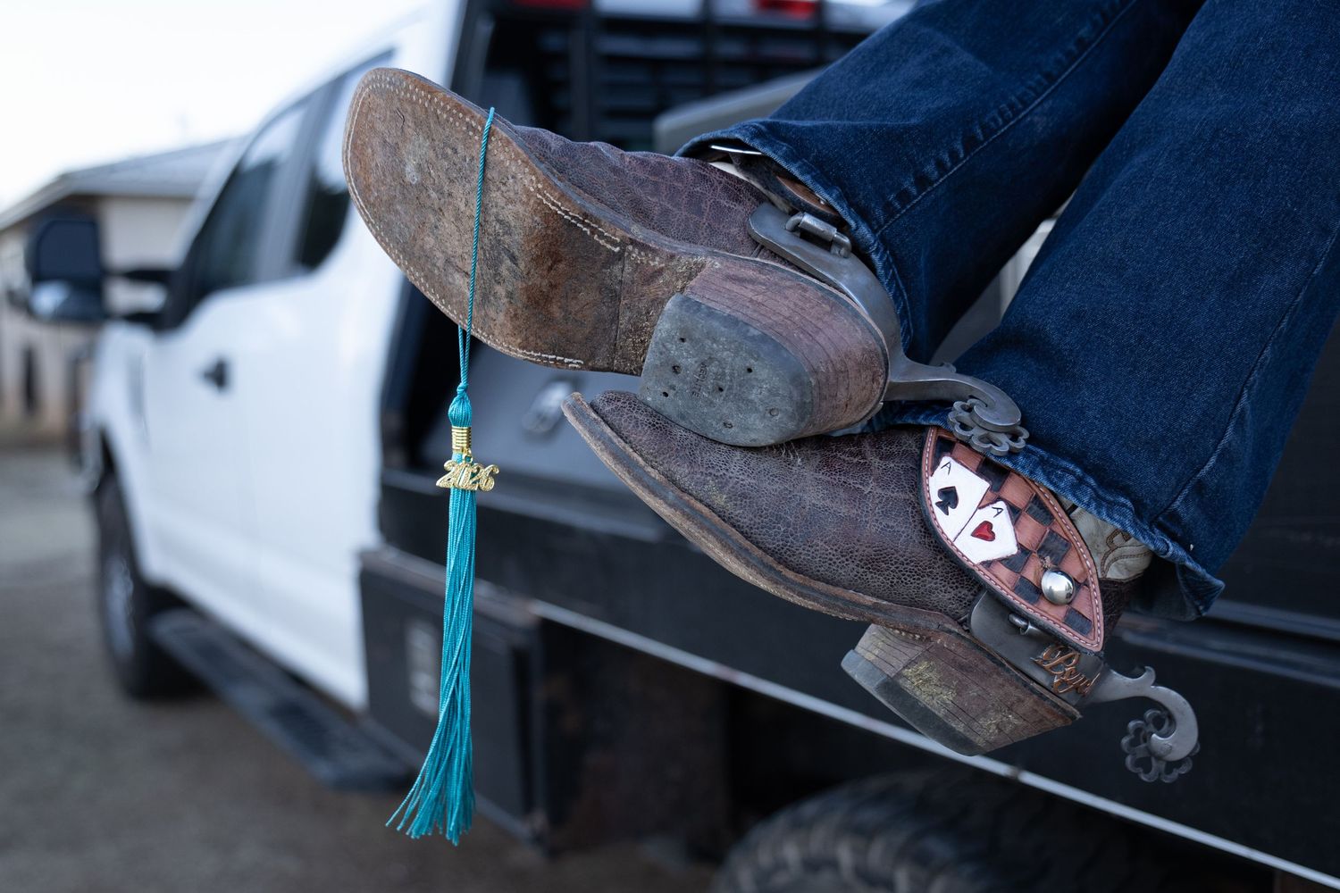 Close-up of cowboy boots with spurs and turquoise graduation tassel resting on truck tailgate.
