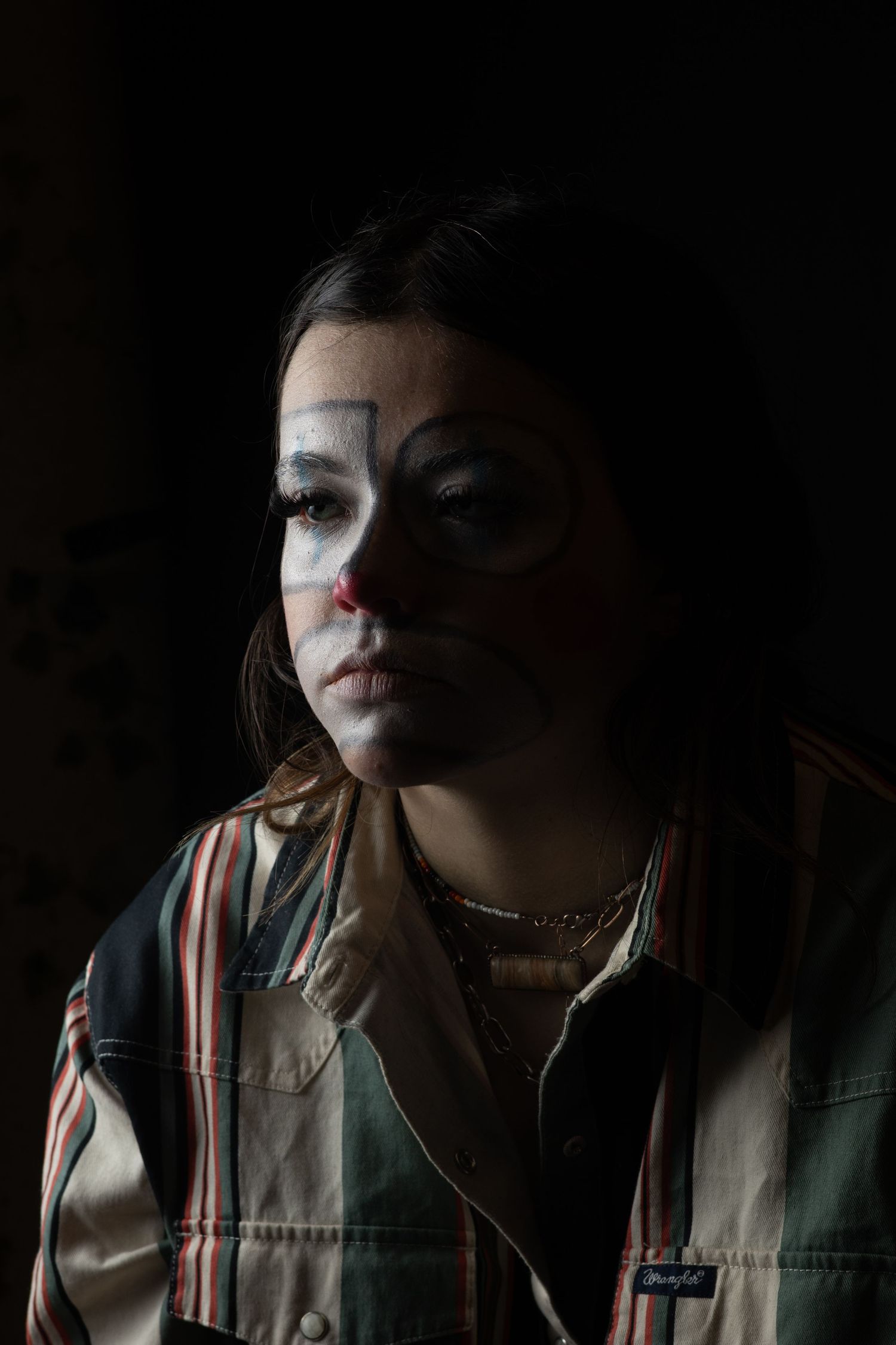 Young woman with dramatic clown face paint in white and red, wearing a striped jacket against a dark background.