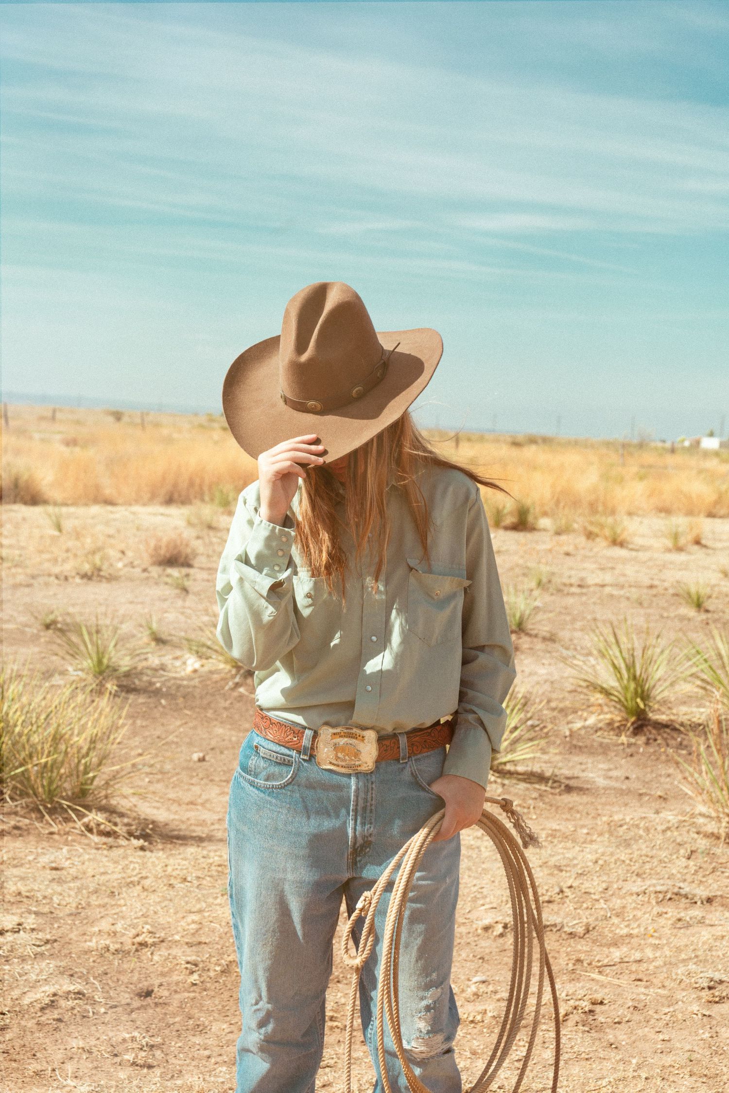 Cowgirl tipping brown felt hat down over face while holding lasso in sun-baked desert landscape.