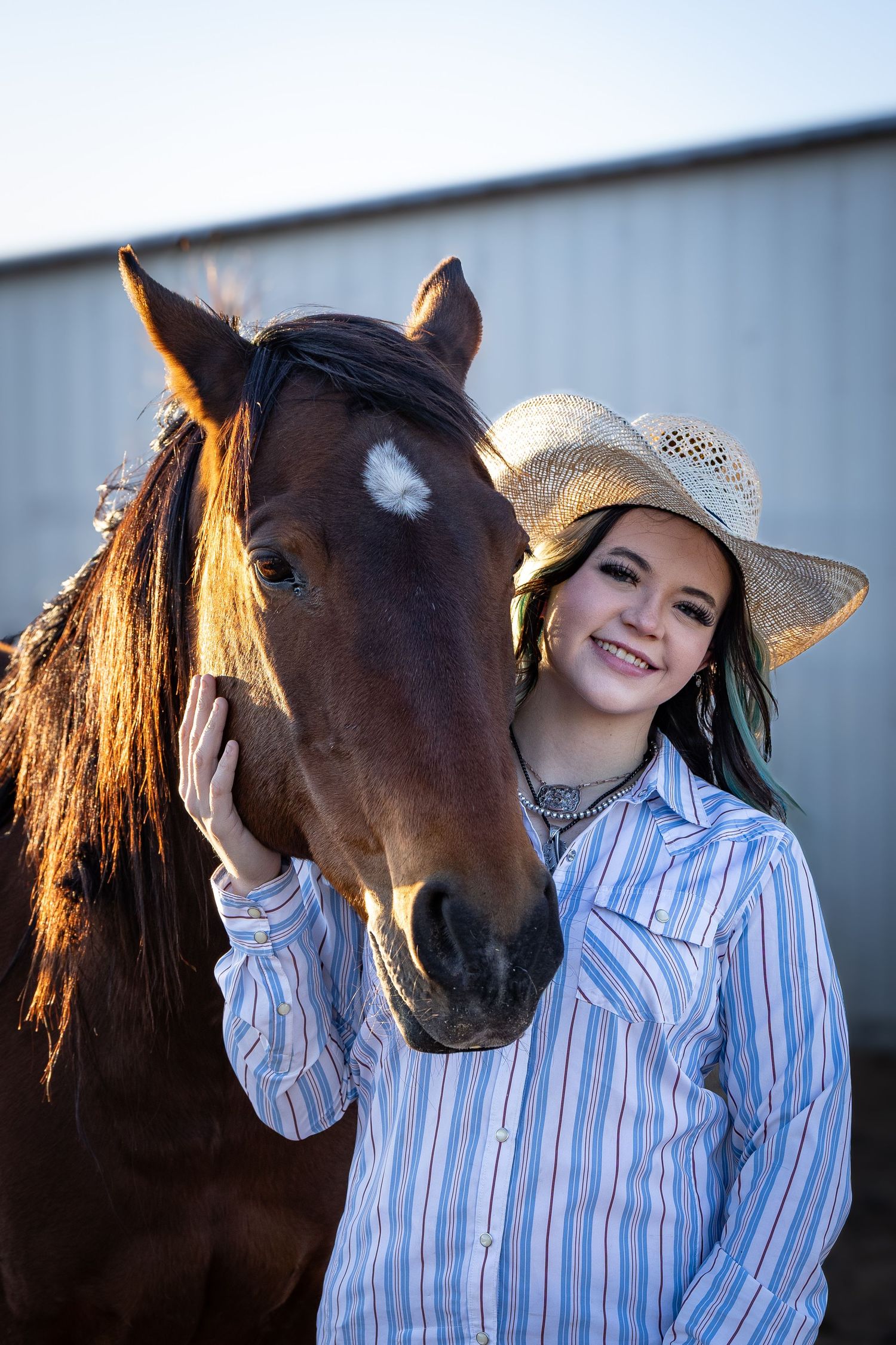 Smiling young woman in cowboy hat and striped western shirt stands beside a brown horse outdoors.