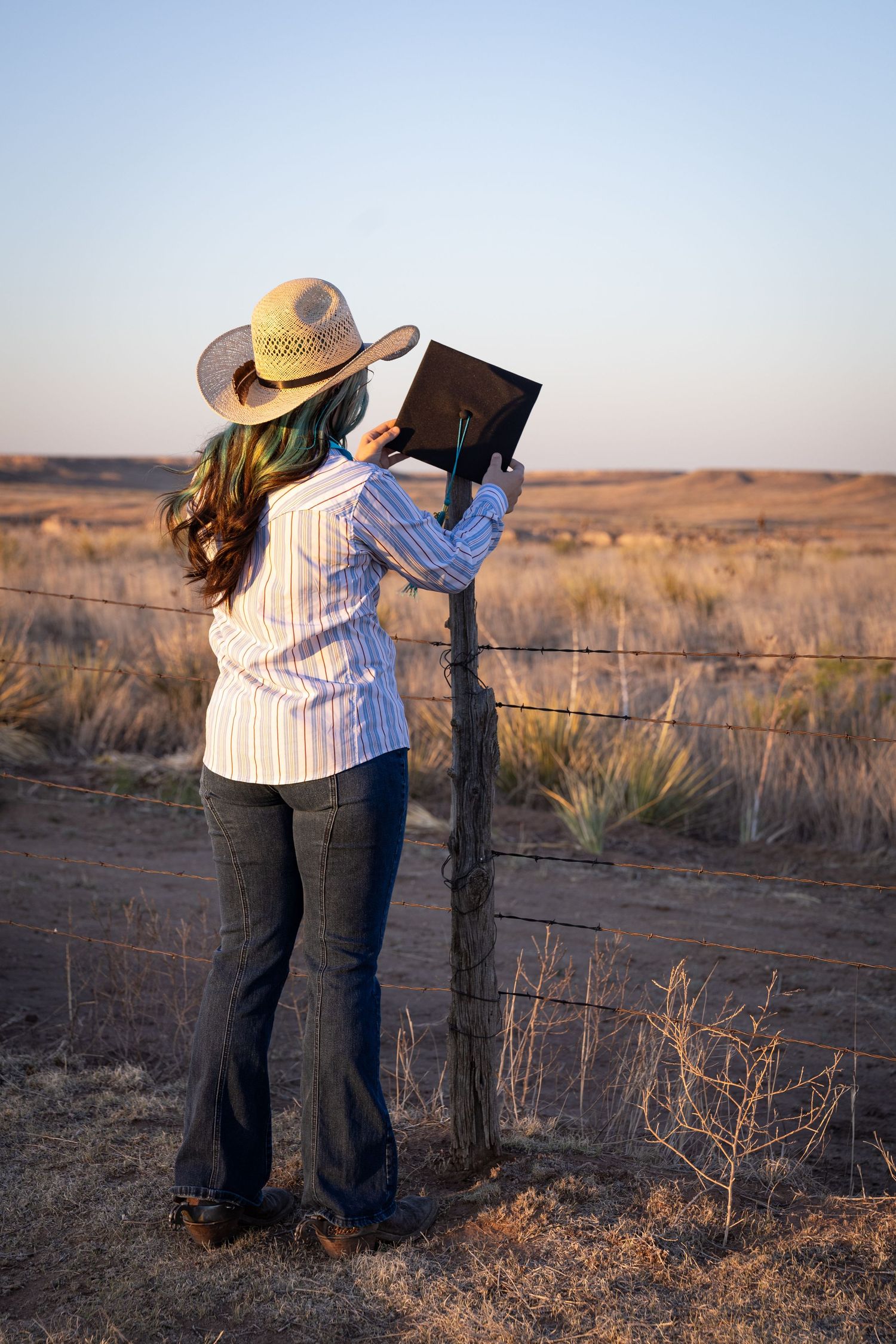 Young woman in cowboy hat and striped shirt holding graduation cap at rustic fence in open prairie at sunset.
