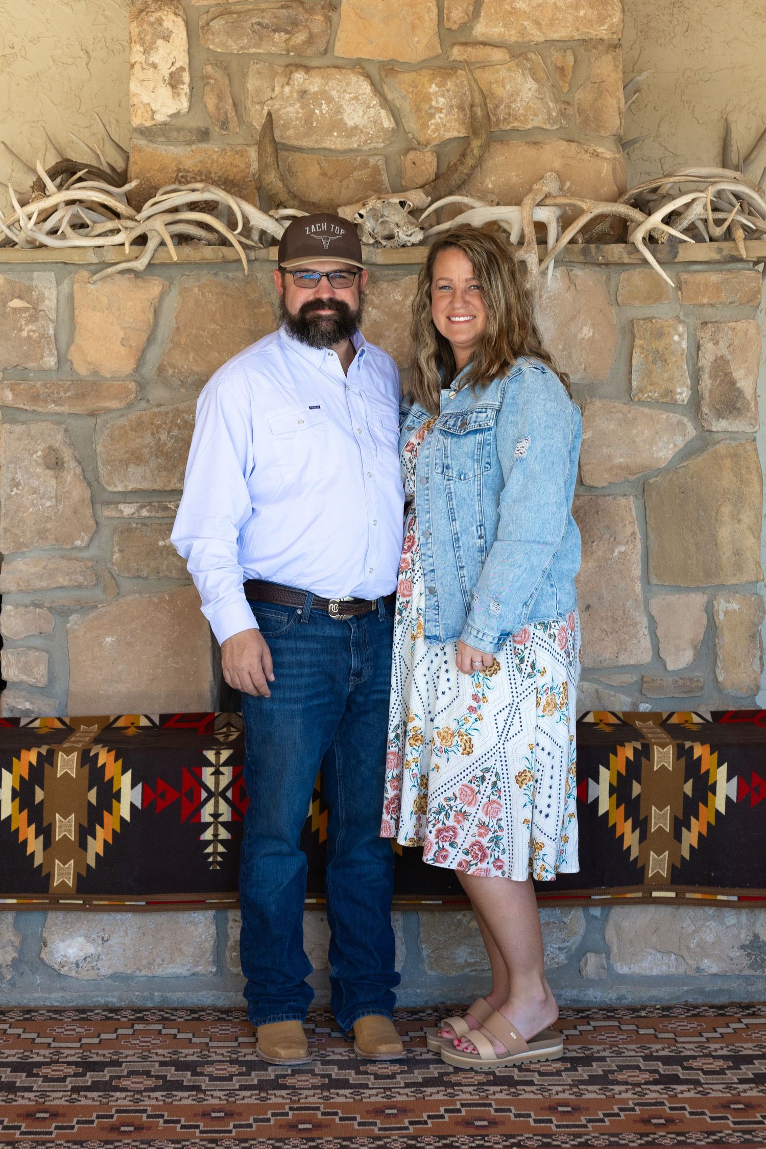 Smiling couple stands together against a rustic stone wall decorated with antlers and a southwestern bench.