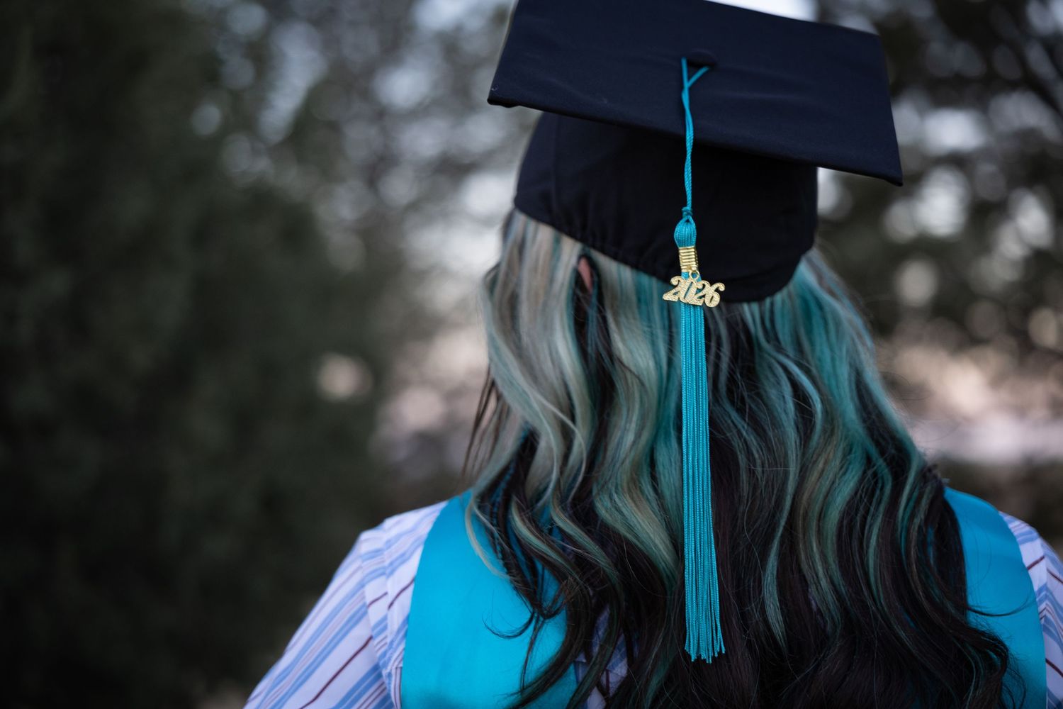 Close-up back view of graduate cap with teal tassel and highlighted wavy hair in outdoor setting.