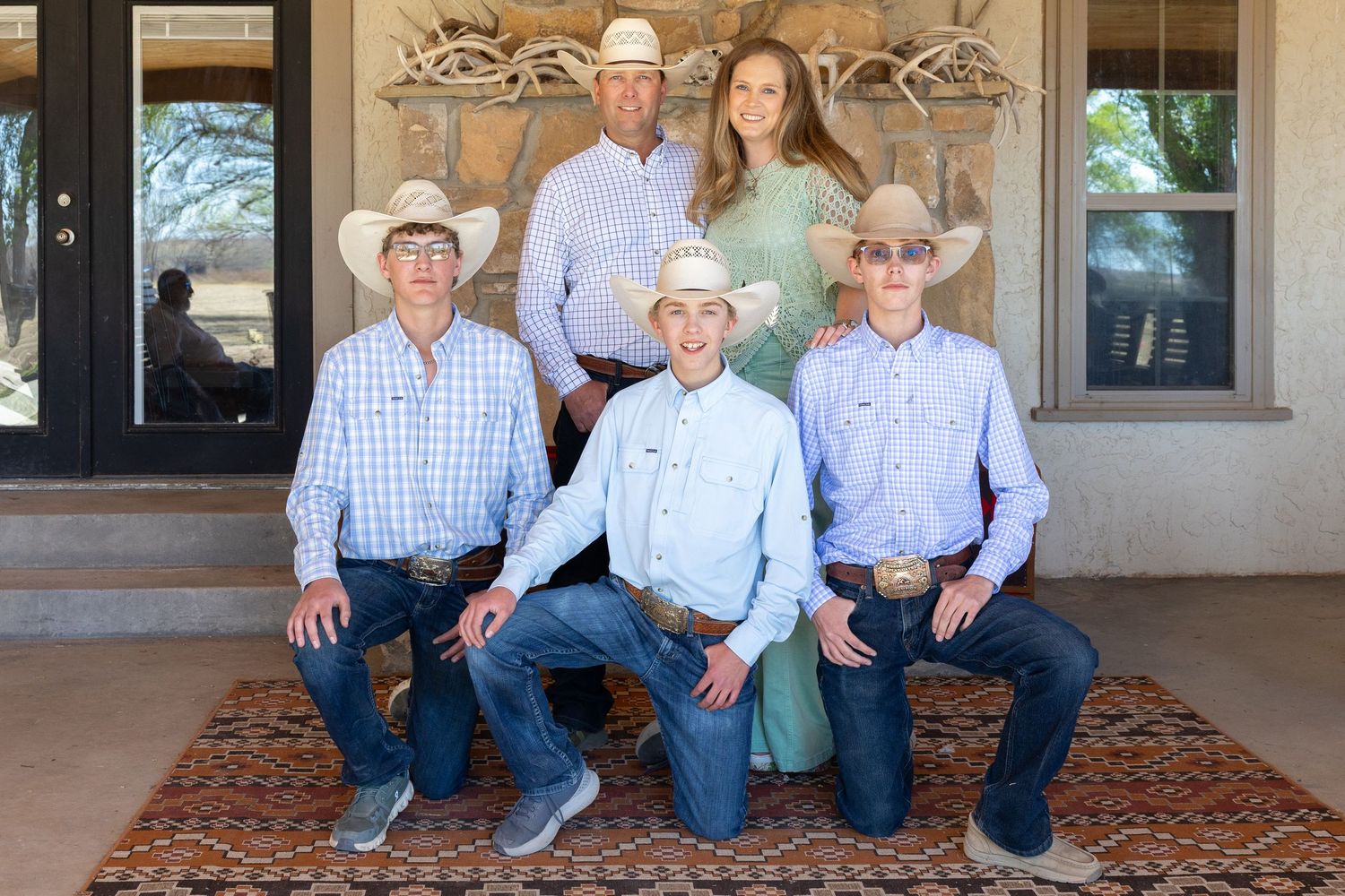 Five family members in cowboy hats and western wear pose on a ranch porch with antler wall decor.