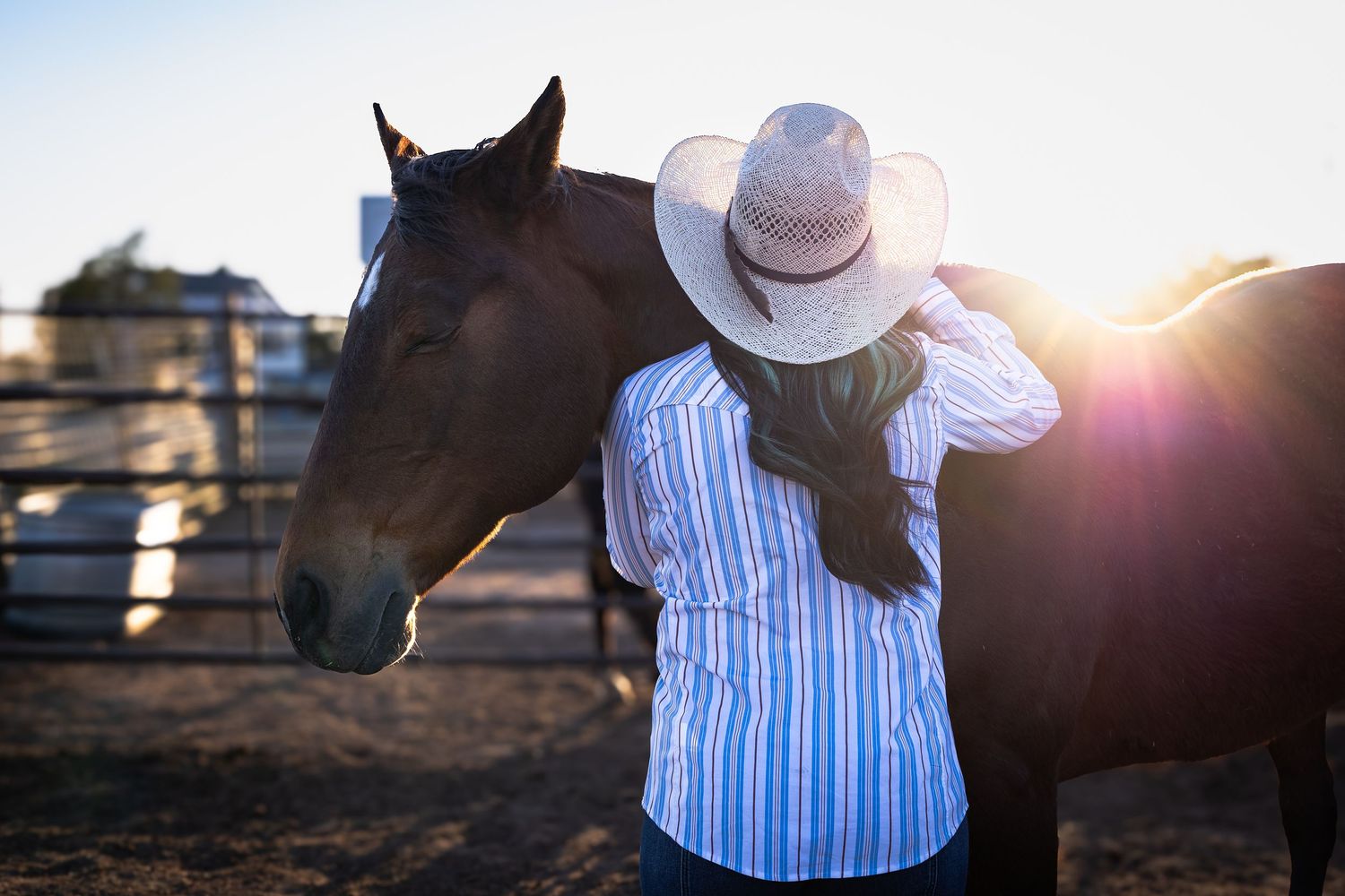 Person in cowboy hat standing beside brown horse at sunset in corral.