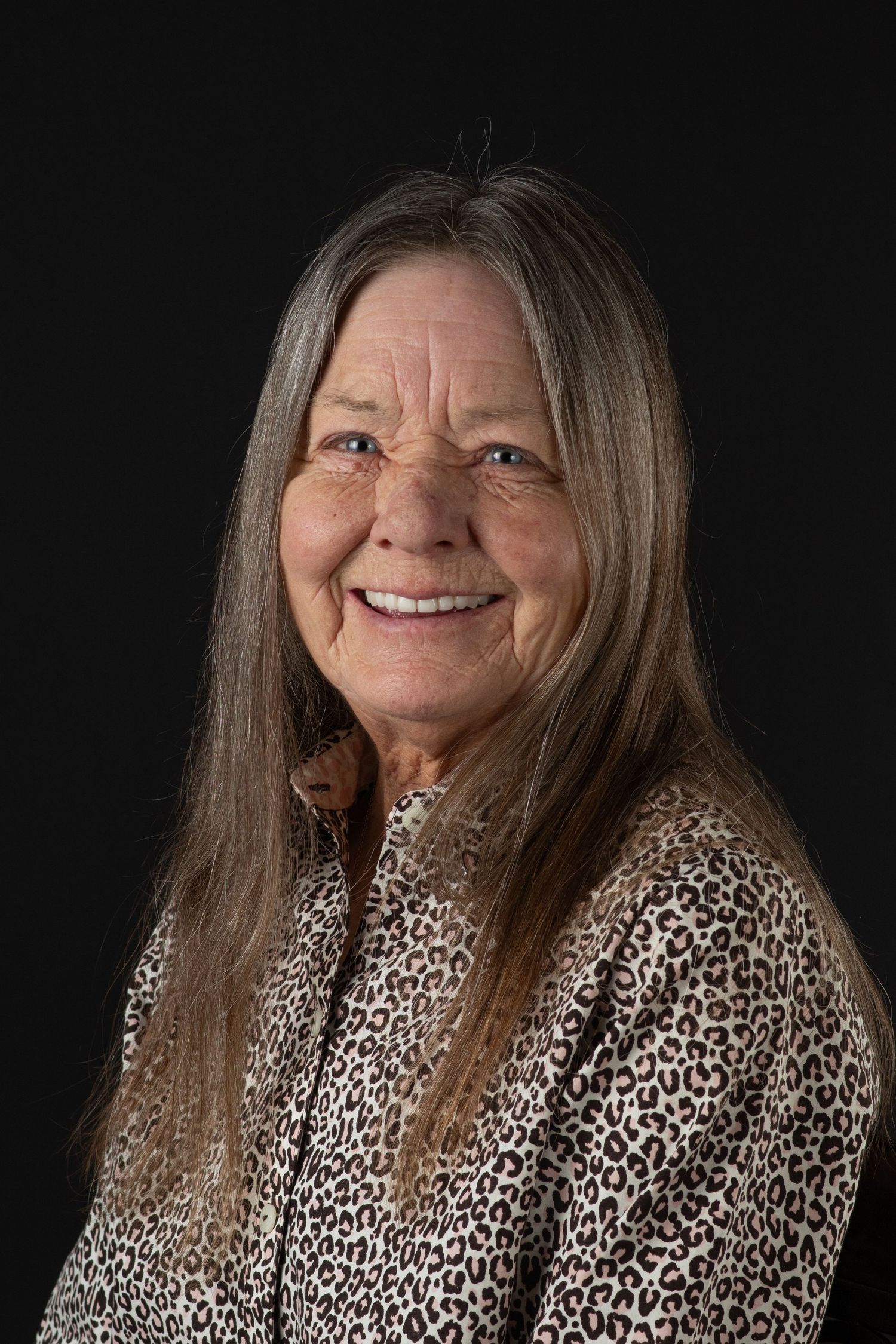 Smiling older woman with long gray hair wearing leopard print top posing against black background.