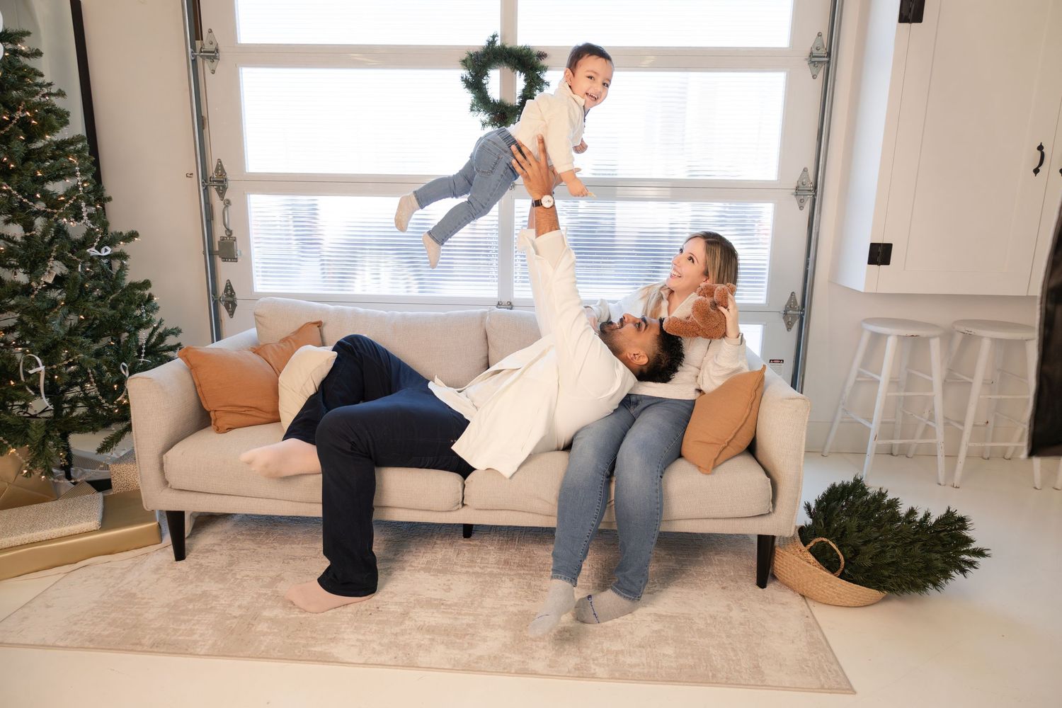 Family plays on sofa near Christmas tree, dad lifting toddler while mom watches in bright living room.