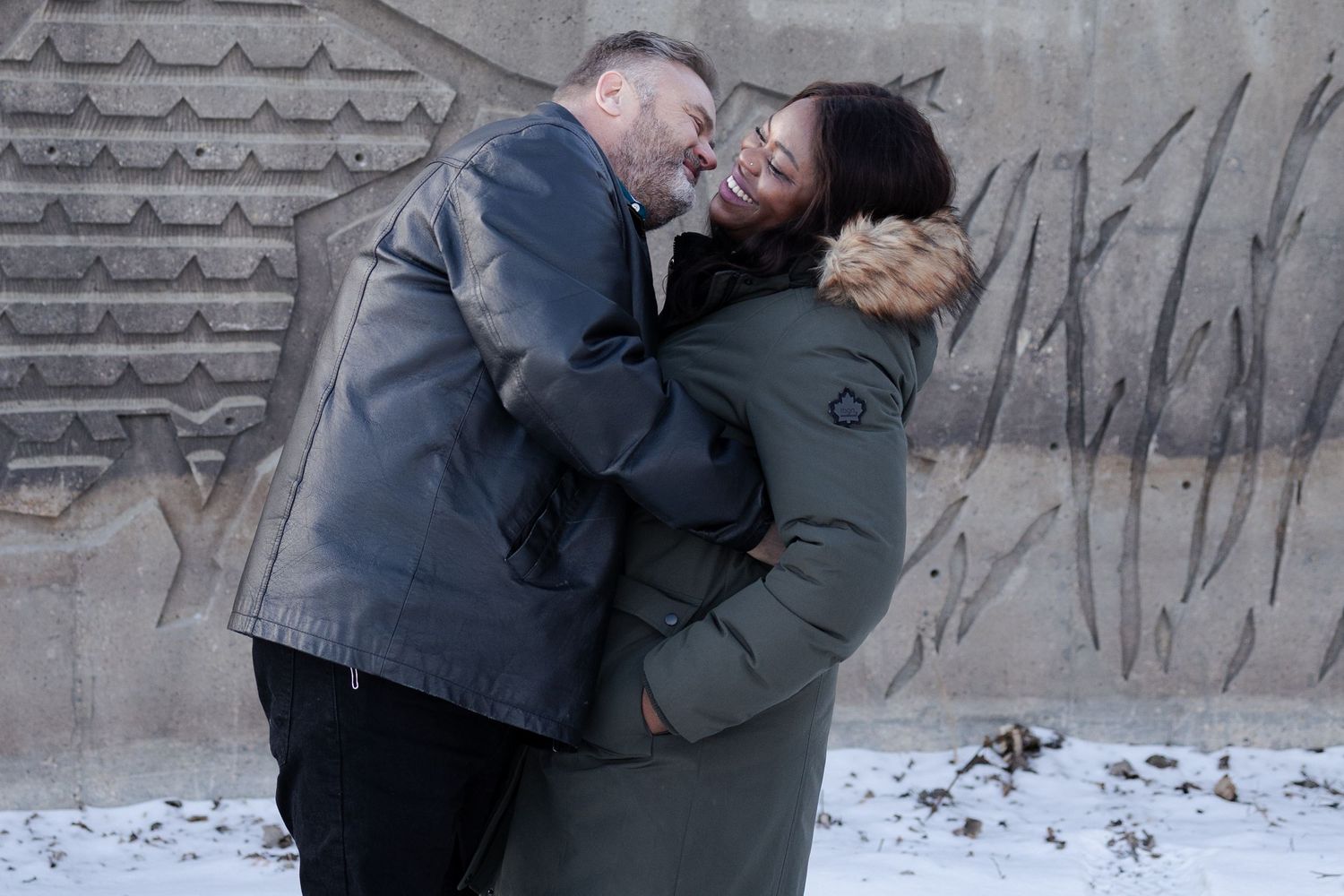 Couple in Winnipeg sharing a playful kiss while embracing outdoors in winter coats near a wall.