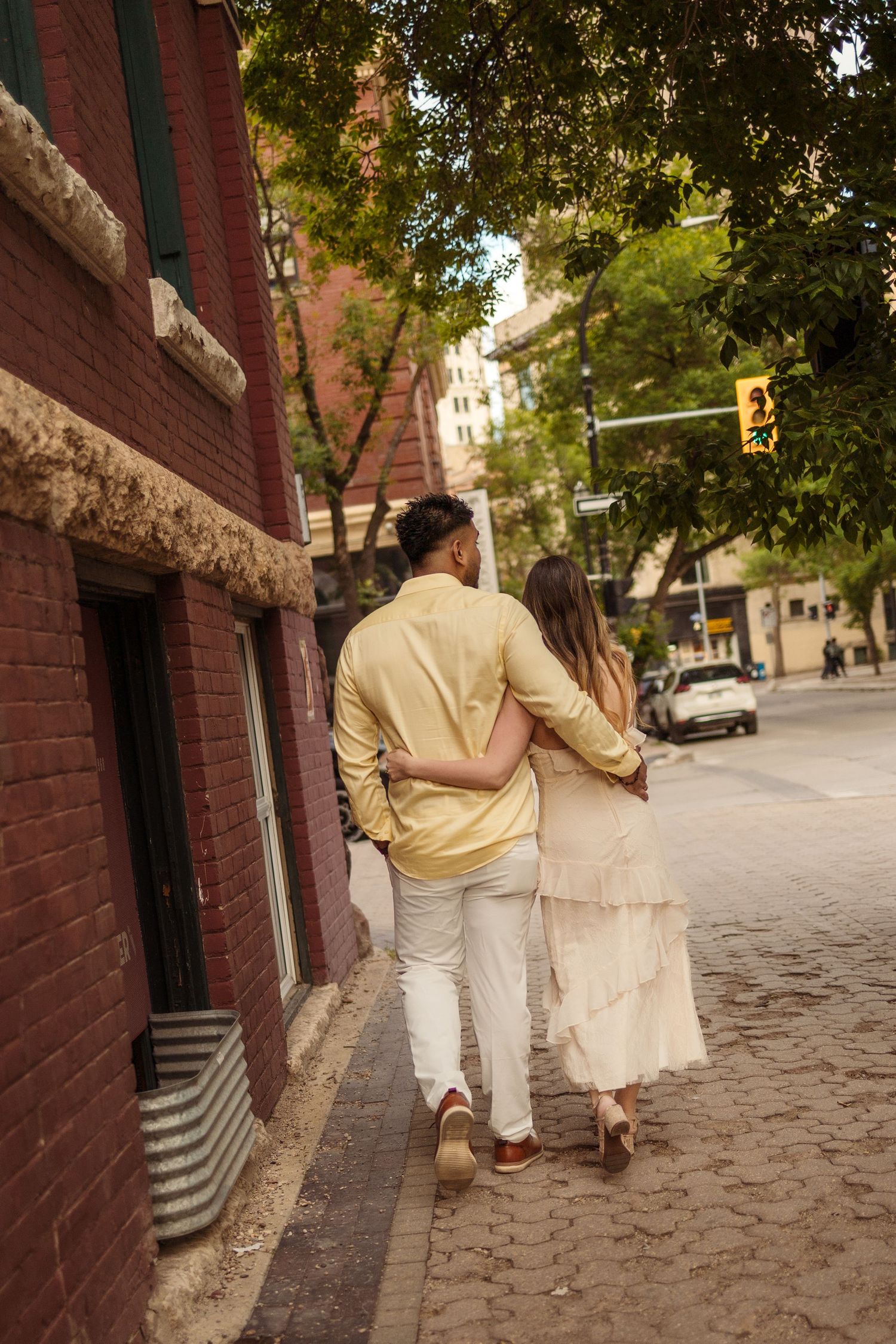 A couple walks arm in arm down a charming brick-lined urban sidewalk, dressed in coordinating cream and yellow outfits.