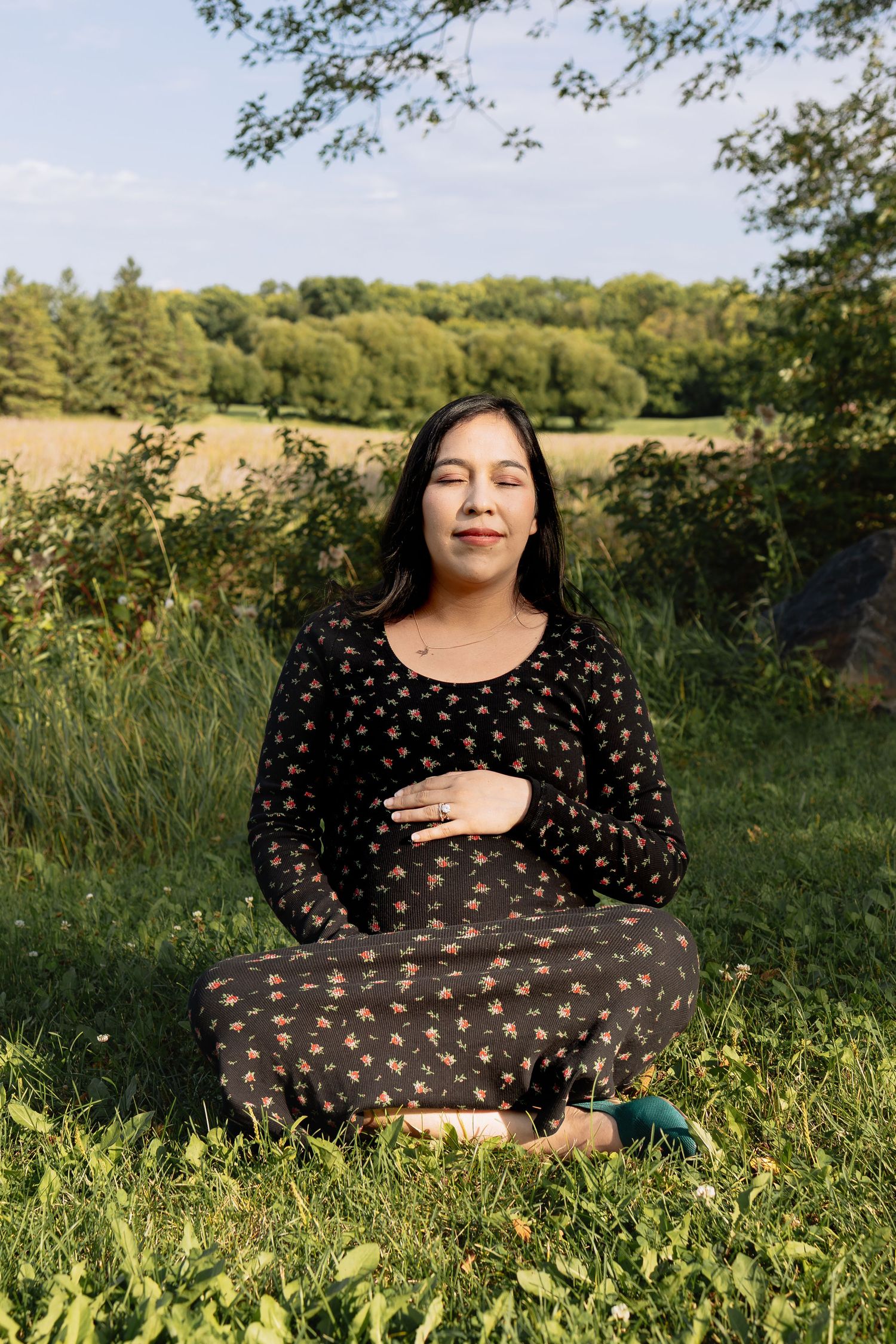 Pregnant woman sitting cross-legged on grass meditating with eyes closed in scenic field in Winnipeg