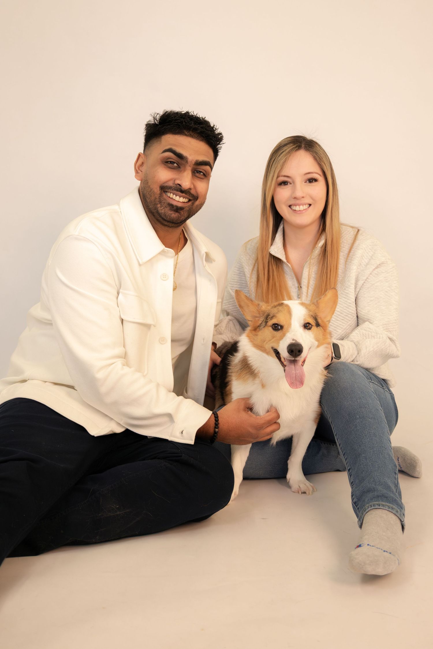 A smiling couple sits on the floor with a happy Corgi dog between them, both dressed in casual neutral-toned outfits.
