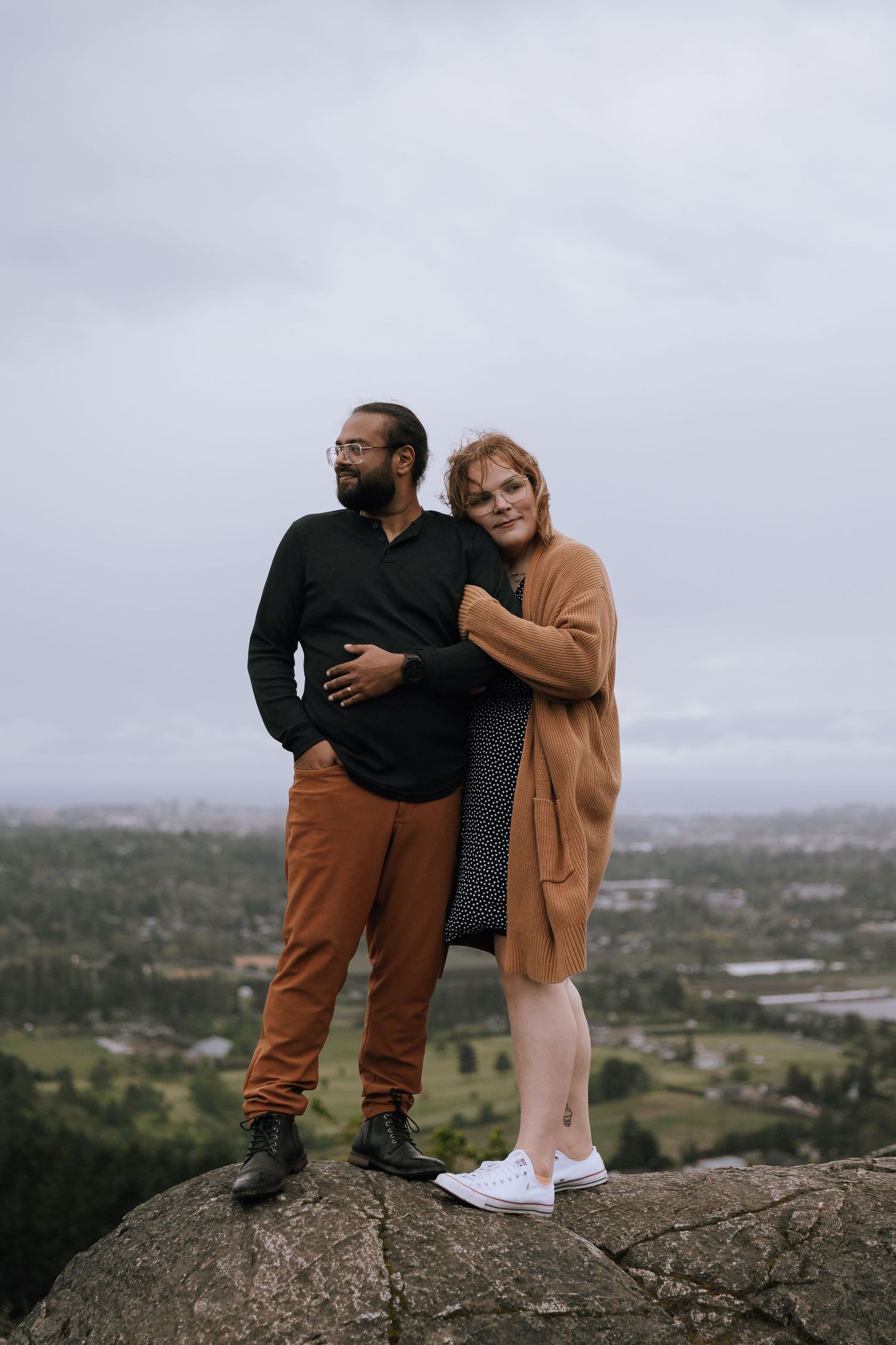 Couple standing on a rocky hilltop embracing with a cloudy cityscape panorama behind them.
