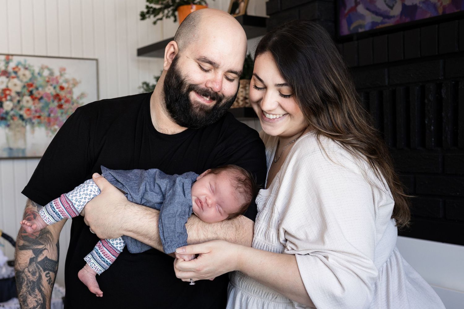 A smiling couple with tattoos lovingly holds their sleeping newborn baby wrapped in a blue blanket indoors.