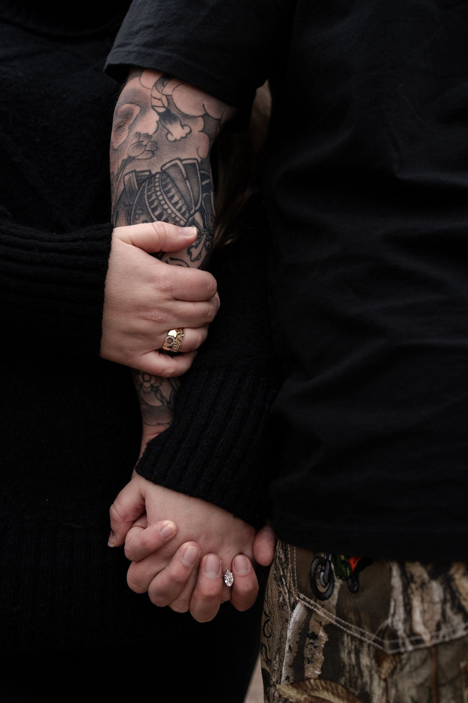Close-up of a couple holding hands, one with a floral tattoo sleeve and engagement ring, wearing black outfits in Winnipeg