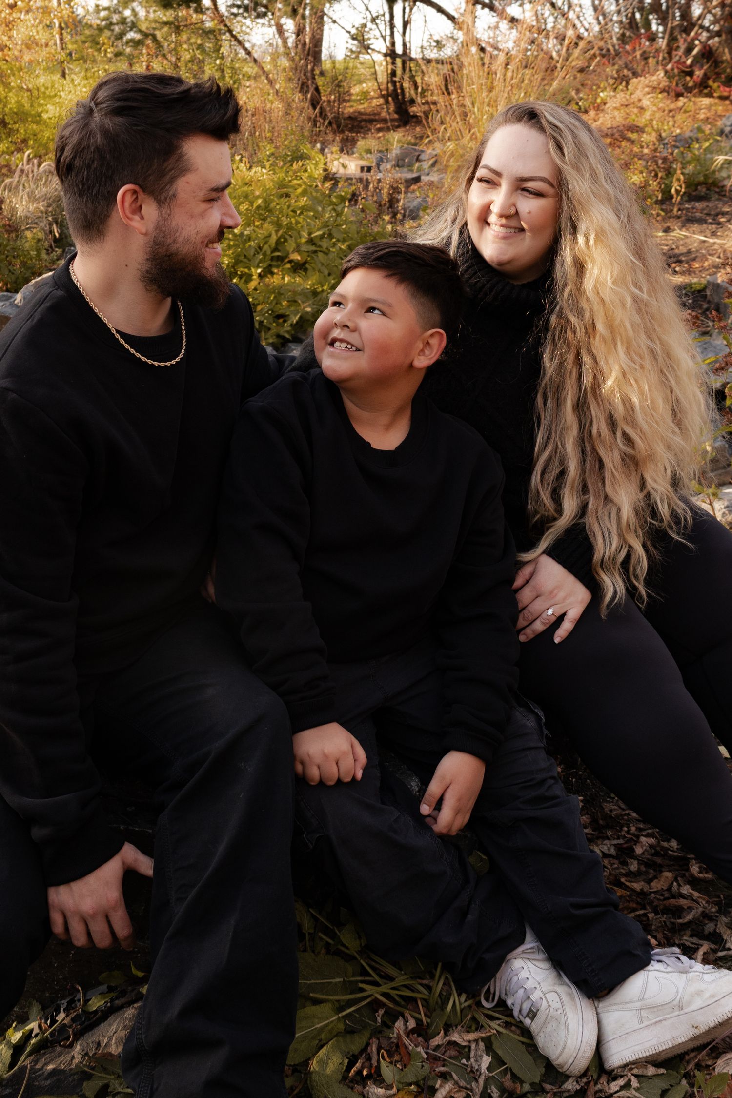 A family of three in black outfits sit together smiling in an autumn outdoor setting in Winnipeg