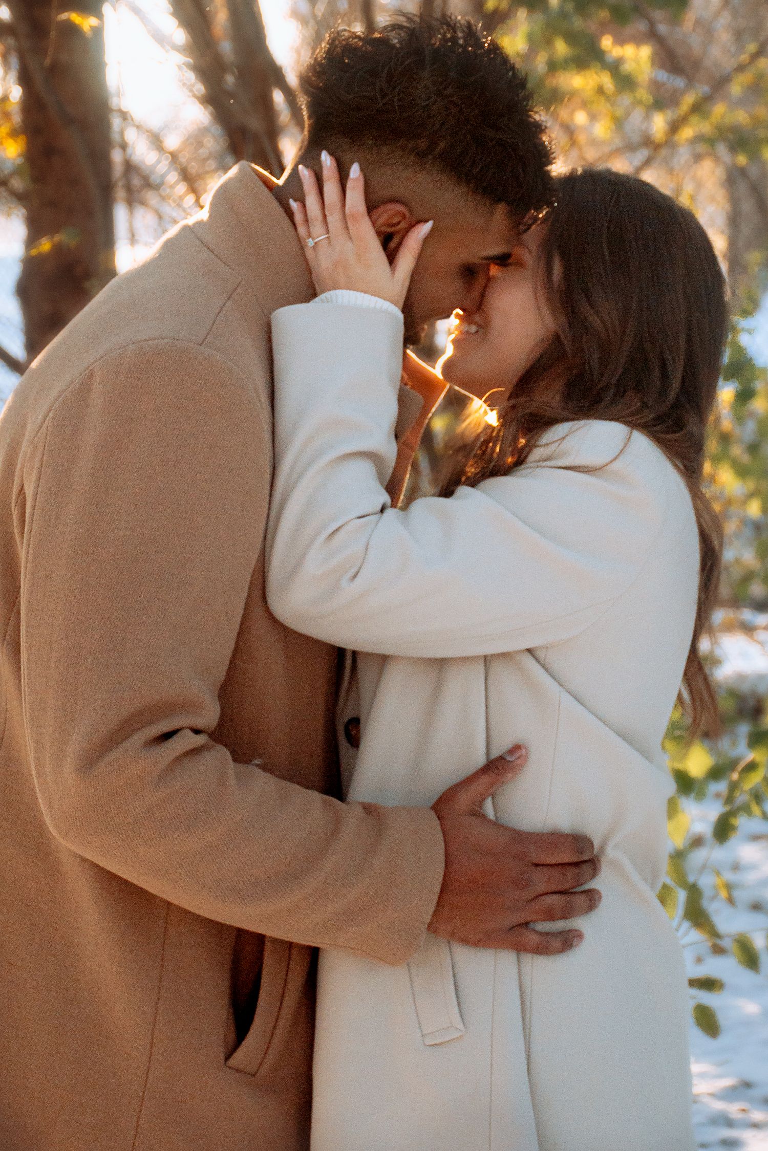 Romantic interracial couple in beige coats embracing and kissing in a sunny winter forest  in Winnipeg