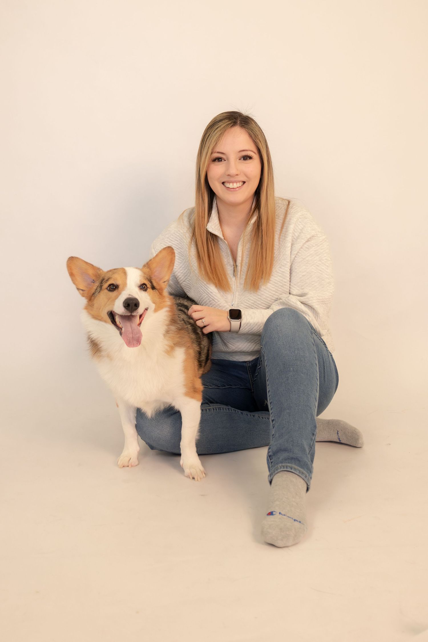 Smiling blonde woman sitting on floor posing with a happy Corgi dog in a studio setting.