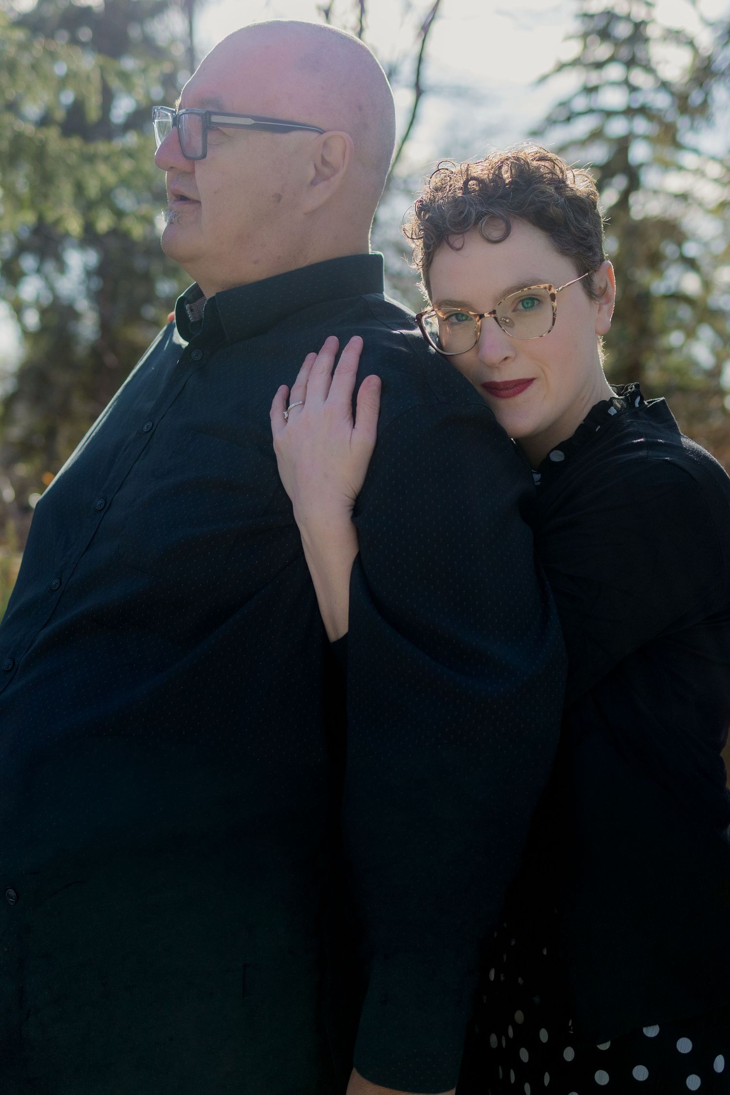 A bald man and short-haired woman in black outfits pose back-to-back outdoors, both wearing glasses.