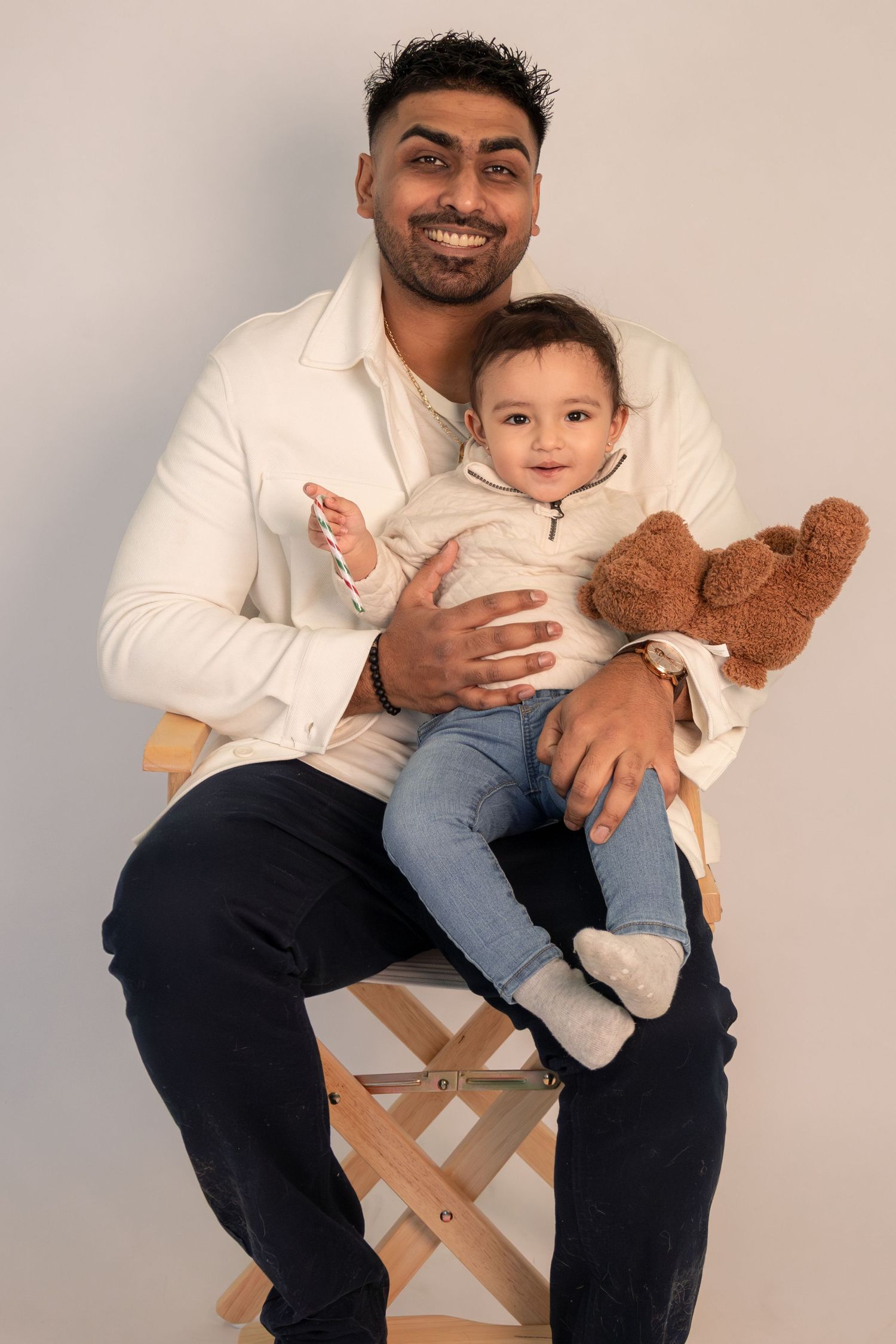 Smiling man with beard holds baby and teddy bear while seated on wooden chair against gray background.