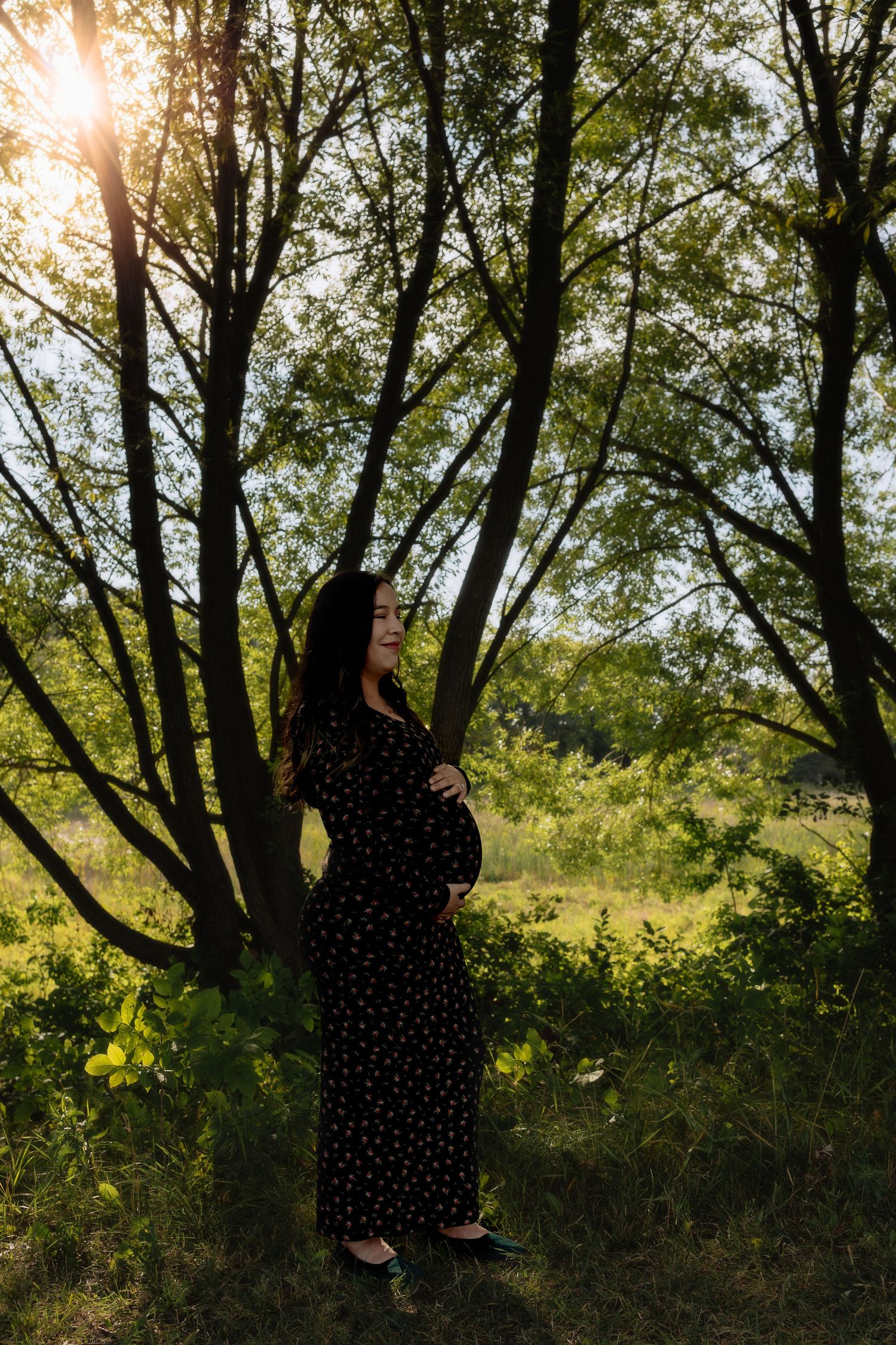 Pregnant woman in black floral dress standing among trees with golden sunlight filtering through leaves in Winnipeg