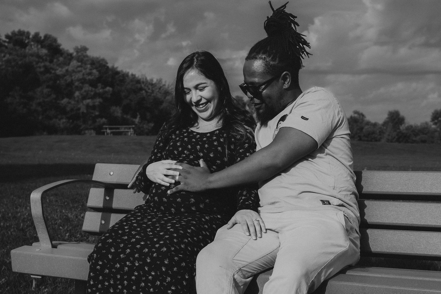 Laughing interracial couple sitting on park bench together enjoying pregnancy moment in black and white in Winnipeg