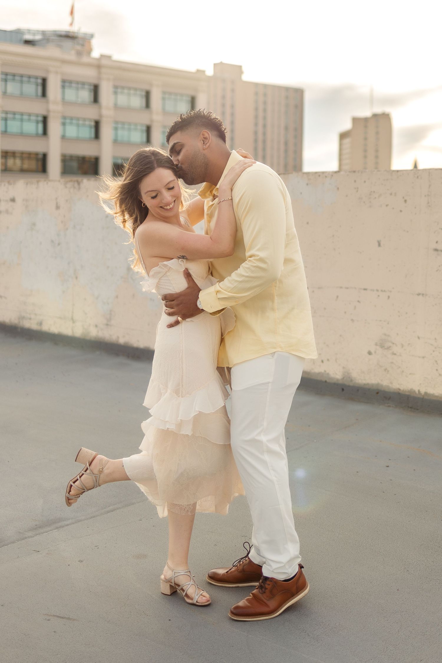 A couple shares a romantic embrace on a rooftop, the man in yellow kissing the woman in a ruffled cream dress at golden hour.