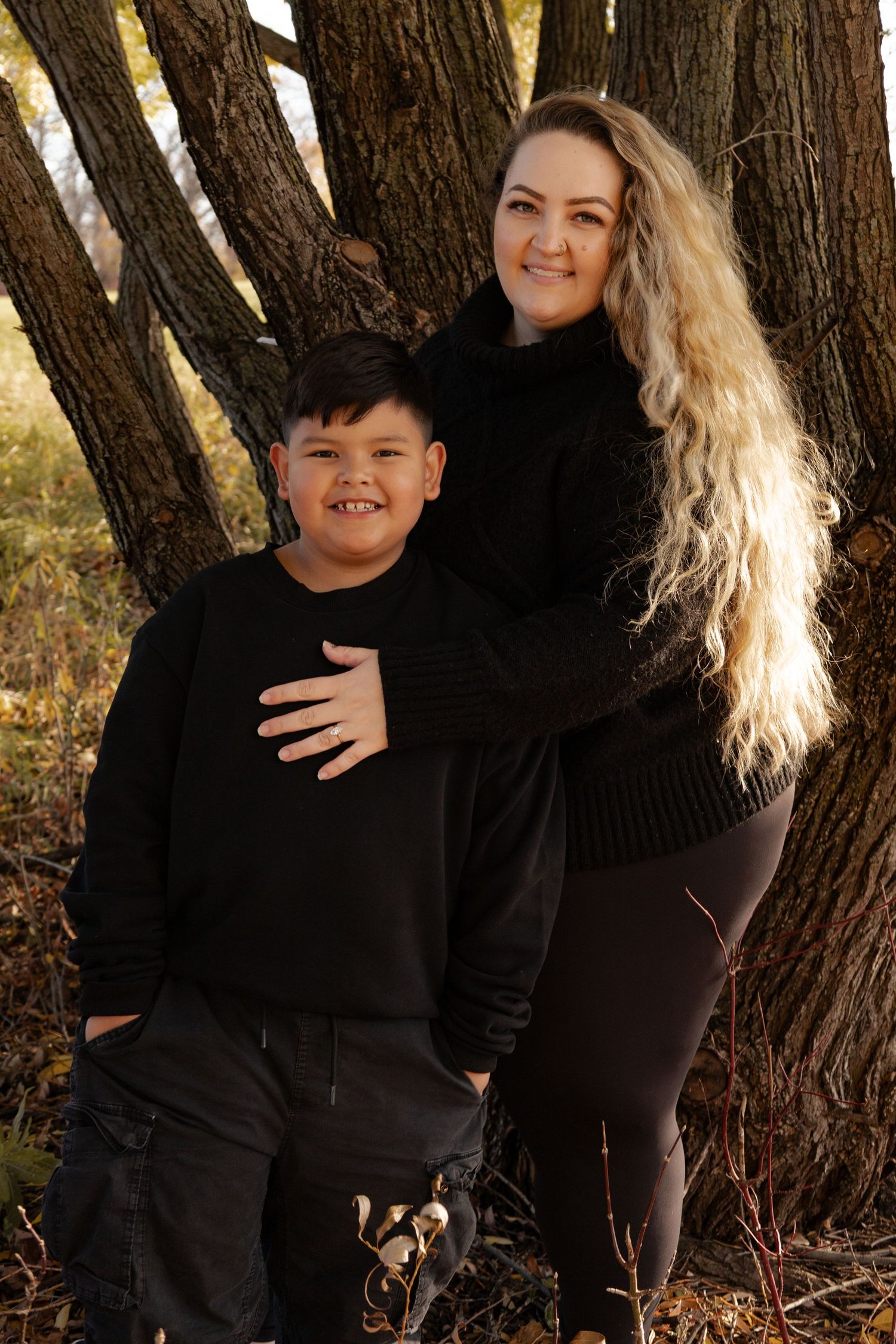 Mother and son smiling together against a textured tree trunk in a fall outdoor portrait in Winnipeg